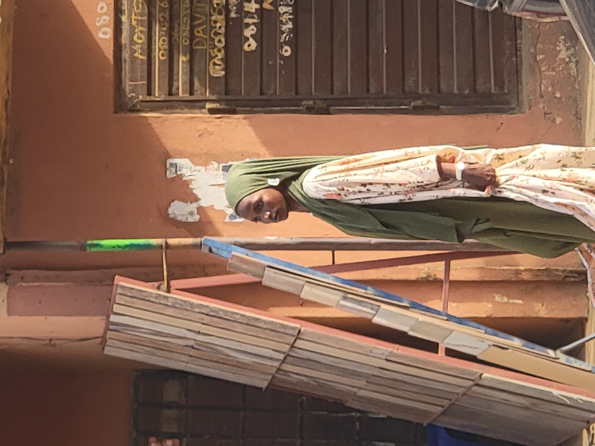 The image appears to be taken outdoors in Maiburiji, Nigeria. A woman wearing a green hijab and a floral-patterned garment is the central subject. She is positioned in front of a building with a boarded-up window. In the foreground, there is a structure that appears to be made of stacked wooden planks with blue accents. A green object is visible behind this structure. The building behind the woman has an orange exterior and shows signs of age and wear. The boarded-up window has text written on it. The lighting indicates that the image was taken in daylight.