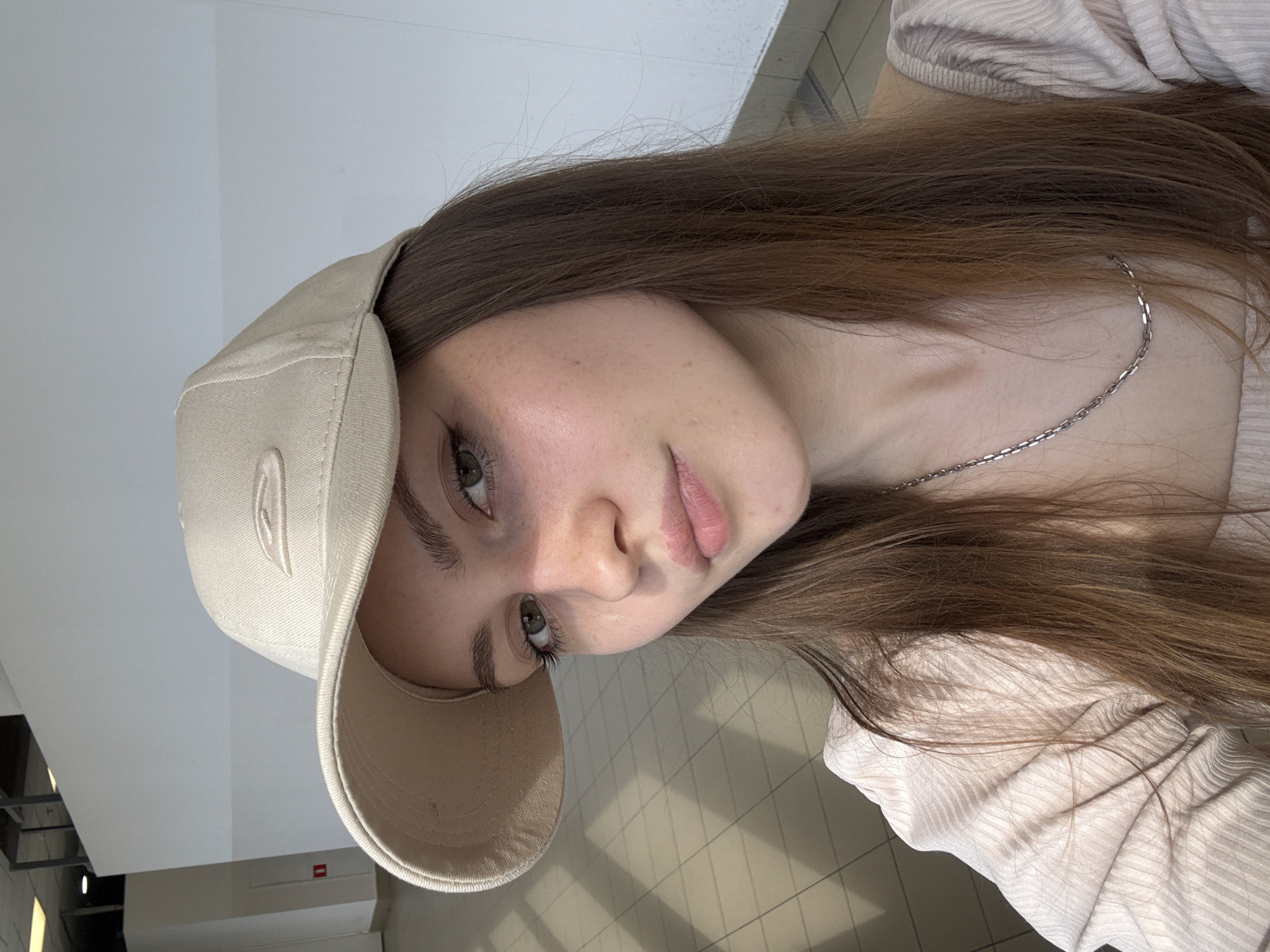 The image is a close-up portrait, oriented ninety degrees clockwise, featuring a young individual, likely female, with fair skin, long straight brown hair, and light-colored eyes that appear green or hazel. Her head is tilted, and she looks directly at the camera with a neutral, slightly contemplative expression.
She is wearing a light beige baseball cap, worn somewhat casually, and a thin, silver-toned chain necklace. Her top is a light, possibly beige or pale pink, long-sleeved garment with a discernible ribbed texture. Her lips are a natural pink color, and she has visible eyelashes and well-defined eyebrows.
The background indicates an indoor setting, possibly a modern public or commercial building. There are light-colored walls and ceiling surfaces with clean architectural lines. In some areas, reflections or structural details suggest glass or polished surfaces, and a patterned floor is partially visible. The lighting is bright and evenly distributed, consistent with a well-lit indoor environment. The context provided places the location in Saint Petersburg, Russia. There is no visible text in the image.