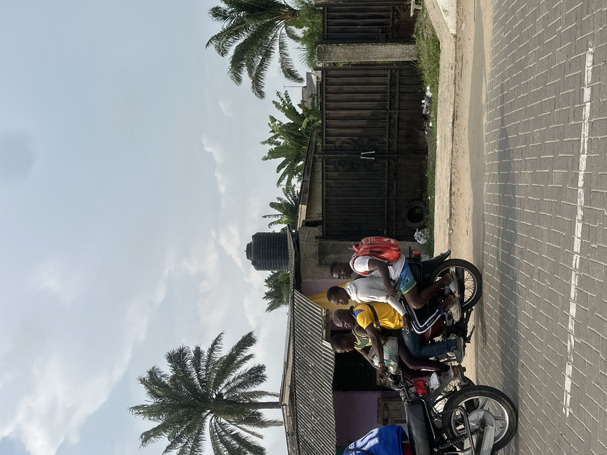 In Badagry, Nigeria, four young males are seated on a motorcycle. The rider, wearing a white t-shirt, is facing forward, while the three passengers are positioned behind him. One passenger in the middle wears a yellow t-shirt, and the passenger at the very back is wearing a blue top. A red backpack is visible on the seat behind the rider. The motorcycle is parked on a paved surface, bordered by a narrow strip of grass and a dark metal gate. Beyond the gate, palm trees and other lush vegetation are visible. The sky is partially cloudy.