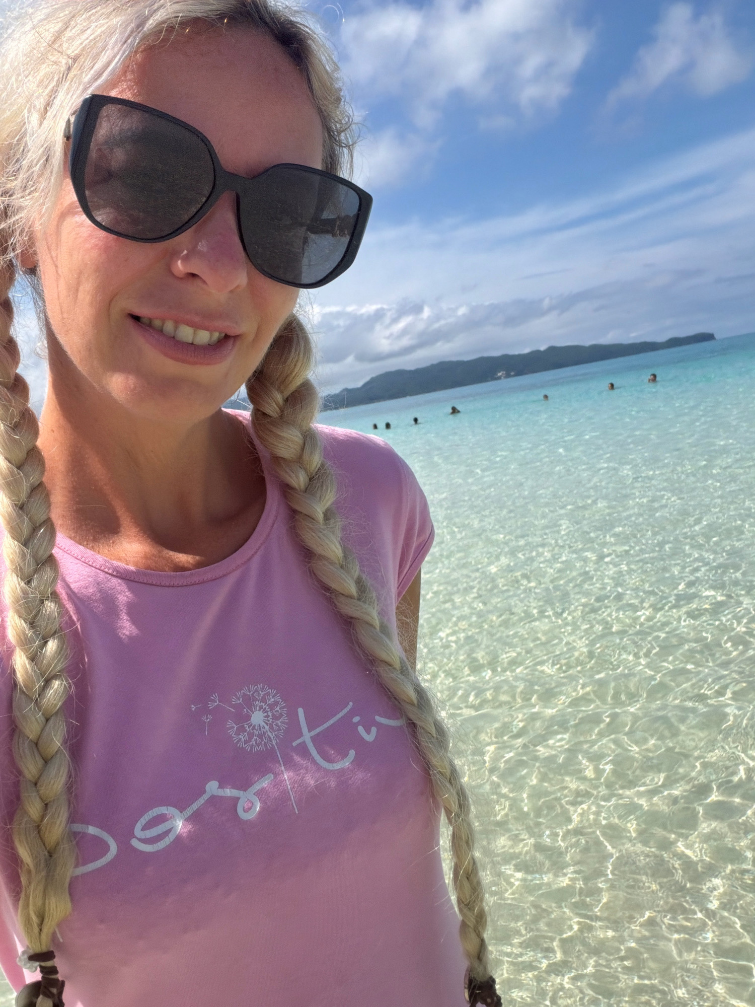 A blonde woman with braided hair and large sunglasses is taking a selfie on a beach. She is wearing a pink t-shirt with a white dandelion graphic and text. The ocean is clear and turquoise, with shallow waves and a sandy bottom visible. In the background, several people are in the water, and a lush green island with buildings is visible in the distance. The sky is blue with scattered white clouds. This scene is likely located on Boracay Island in Malay, Philippines, known for its pristine white sand beaches and crystal-clear waters.