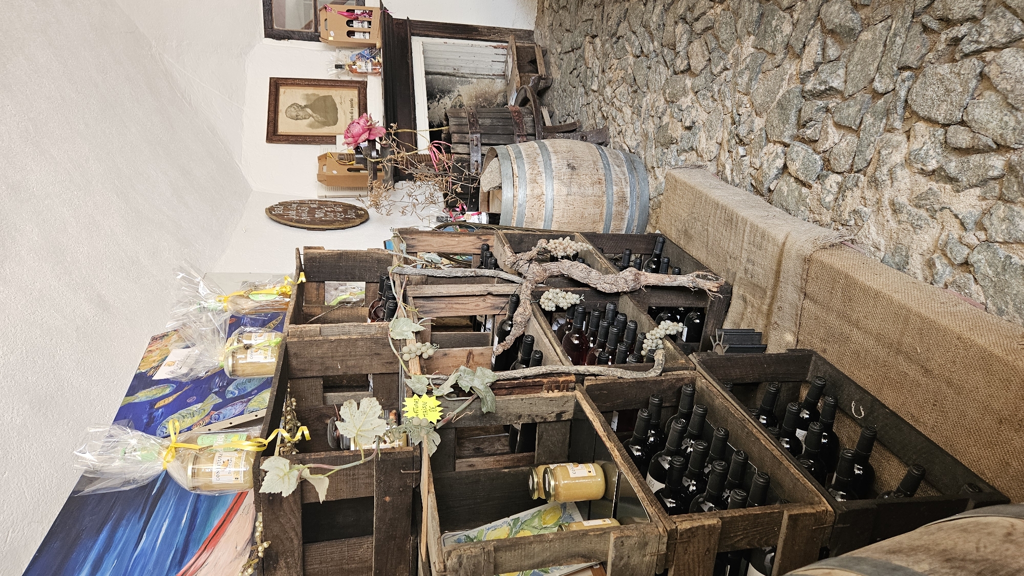 The image depicts the interior of a rustic shop, likely a wine or specialty food store, with a cobblestone floor and exposed wooden crates used for display. On the left, a multi-tiered wooden shelving unit is filled with numerous bottles of wine, arranged horizontally. Decorative elements like artificial grapevines and leaves are intertwined with the shelving and bottles. In the upper left, several jars wrapped in cellophane with yellow ribbons are displayed on top of the crates, alongside a colorful painting on the wall.
Towards the center and right of the image, a large wooden barrel stands on the floor. Behind it, a fireplace with a dark mantelpiece is visible. Above the fireplace, a framed portrait of a man is hung. Further to the right, there are more bottles, some displayed in boxes, and a pink flower. The overall impression is one of a charming, traditional establishment, possibly located in a historic area. While the prompt mentions Sant'Antonino, France, this specific location cannot be confirmed from the visual information alone.