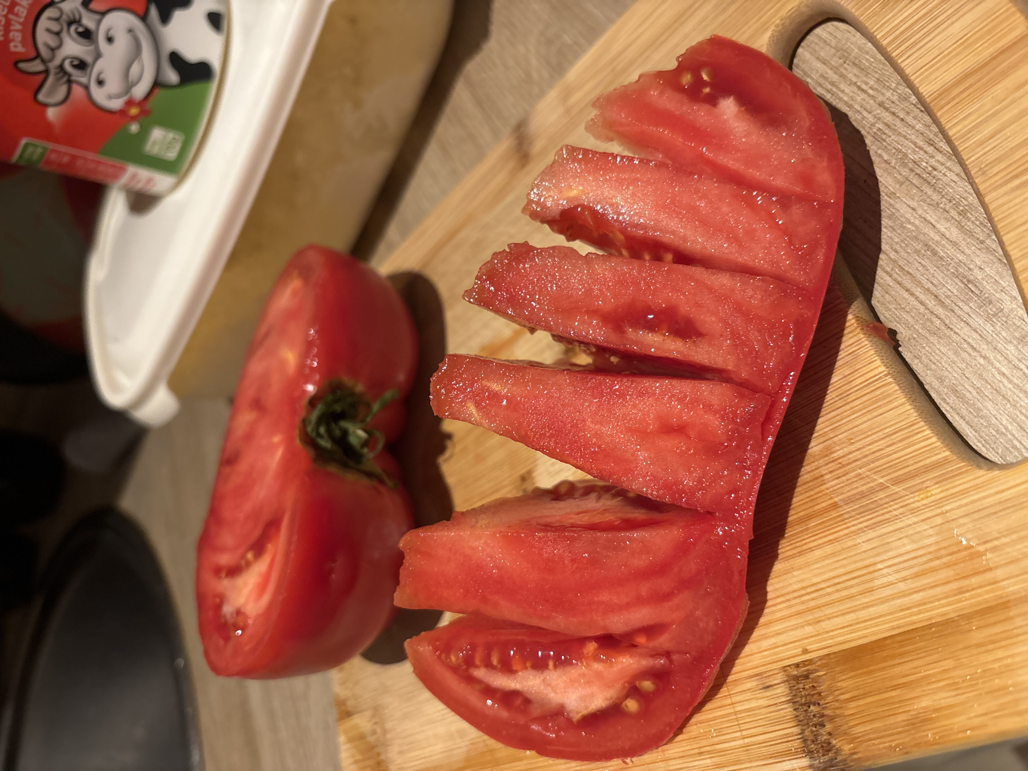 The image depicts a wooden cutting board with a halved tomato, one half sliced into segments. Next to the board is a container with an image of a cow on its label. The overall scene appears to be indoors. There is no evidence of location or interaction with people.