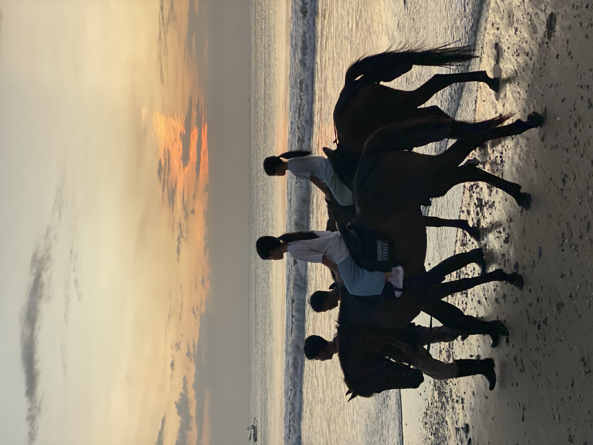 At sunset on Kuta Selatan beach in Indonesia, two young girls are riding horses along the shoreline. They are silhouetted against the warm glow of the setting sun, with the ocean waves gently lapping at the sand. Two men, likely guides, are walking beside and behind the horses, assisting with the ride. One of the girls is wearing a white t-shirt and light blue pants, while the other is wearing a similar outfit. The horses are dark brown, and their manes and tails are flowing in the breeze. The sand is wet and reflects the colors of the sky, creating a serene and picturesque scene. In the distance, a small boat can be seen on the water. The sky is painted with hues of orange, pink, and yellow, with scattered clouds adding depth and texture to the horizon. The overall atmosphere is peaceful and tranquil, capturing a moment of natural beauty and human interaction.