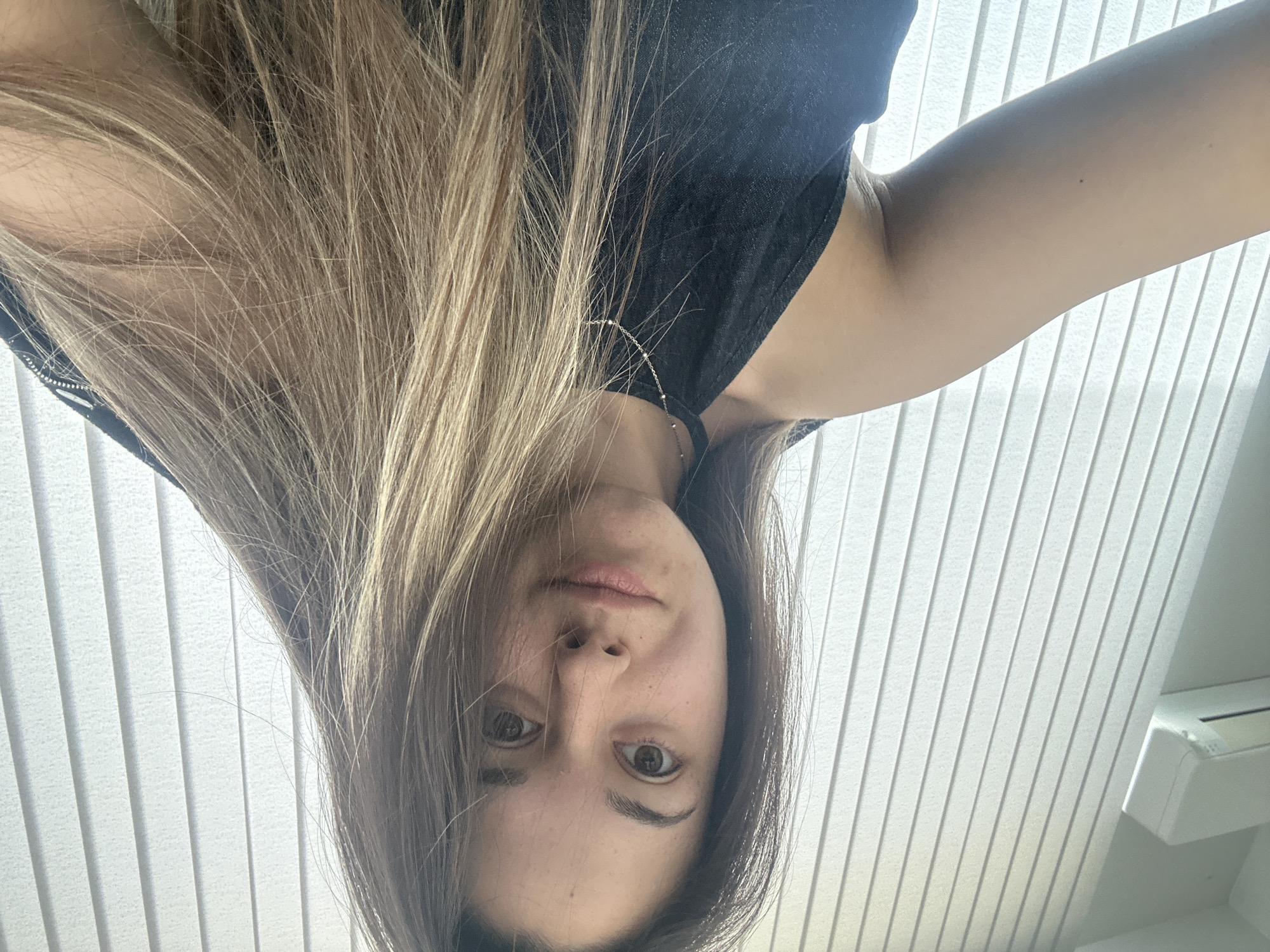 A woman with long blonde hair is holding a camera above her head, pointed downward, capturing a selfie. She is wearing a black tank top and a delicate silver necklace. The background consists of white vertical blinds. A white air conditioning unit is visible in the top right corner of the image. There is no indication that the location is Sofia, Bulgaria.