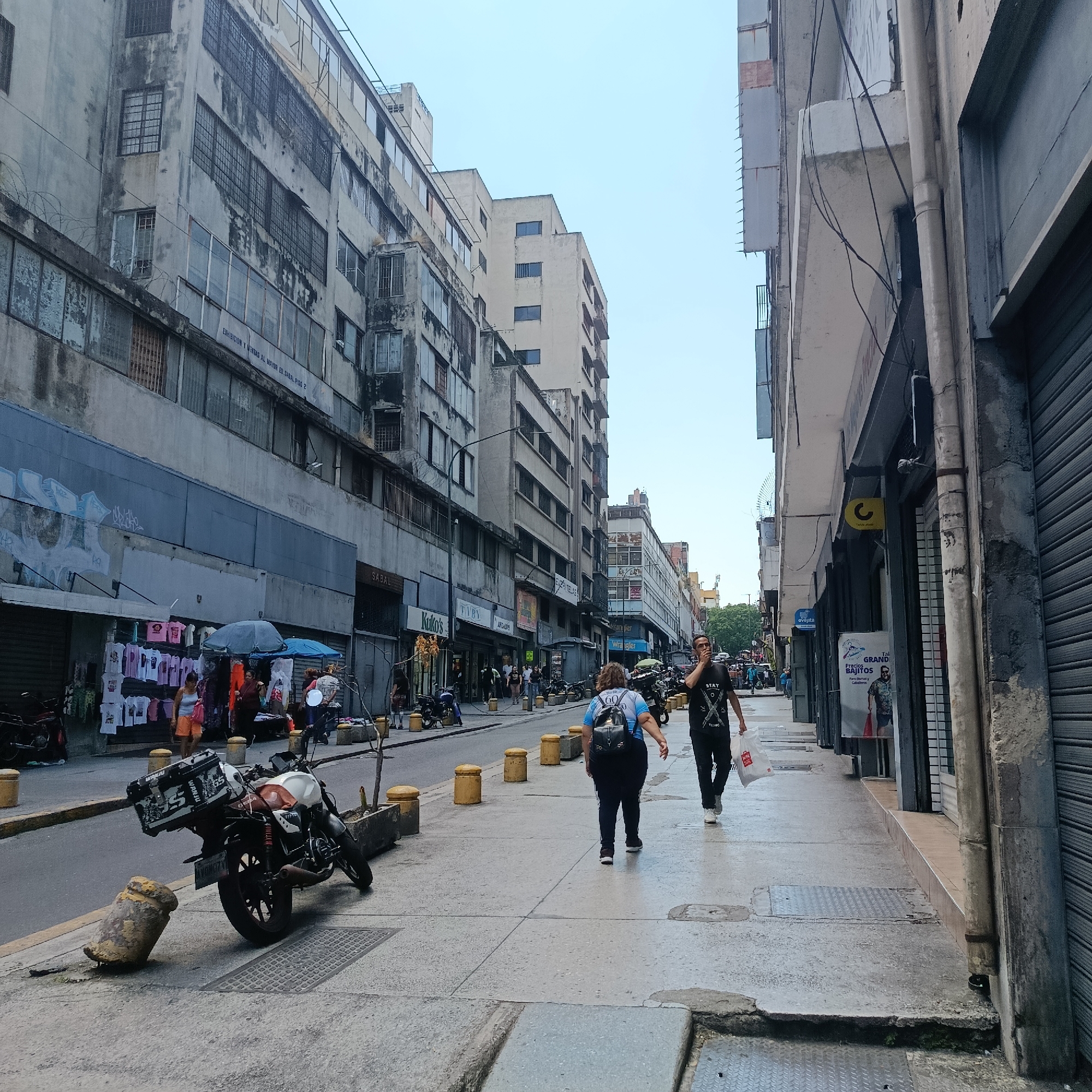 The image depicts a daytime street scene on a commercial thoroughfare in a central district of Caracas, Venezuela, with the prominent "Edificio Sabal" visible on the left, indicating a location associated with wholesale businesses.
The environment is characterized by multi-story concrete buildings lining both sides of the street, appearing aged with visible staining, exposed rebar, and numerous windows, many secured by metal bars or corrugated roller shutters. Graffiti is present on lower building sections on the left. Overhead utility wires are visible. The sky is clear and light blue.
On the left side of the street, multiple storefronts are present, some displaying merchandise like t-shirts on outdoor racks under blue umbrellas, suggesting street vending or open-air retail. A vendor under a light blue umbrella interacts with customers. A woman in a light top and orange shorts stands near one of these displays.
The sidewalk on the right features closed metal roller shutters on most storefronts, with a single visible shop sign bearing a yellow 'C' logo. A tall, thin drainage pipe runs vertically along the building facade.
The street itself is paved, with yellow cylindrical bollards lining the curb, some of which are damaged or fallen. A white and orange motorcycle with a black rear top case (bearing "WRANDU" branding and license plate HXND7X) is parked on the sidewalk on the left, near a fallen yellow bollard. Other motorcycles are parked further down the street.
Approximately 10-15 individuals are present. In the foreground, a person wearing a light blue shirt, dark pants, and a black backpack walks away from the camera on the left sidewalk. To their right, a male in a black t-shirt with a white graphic, dark pants, and white shoes walks towards the camera, holding a white plastic bag and an object to his mouth. Further down the street, other pedestrians are walking in both directions, engaging in commercial activities, or standing near shops.