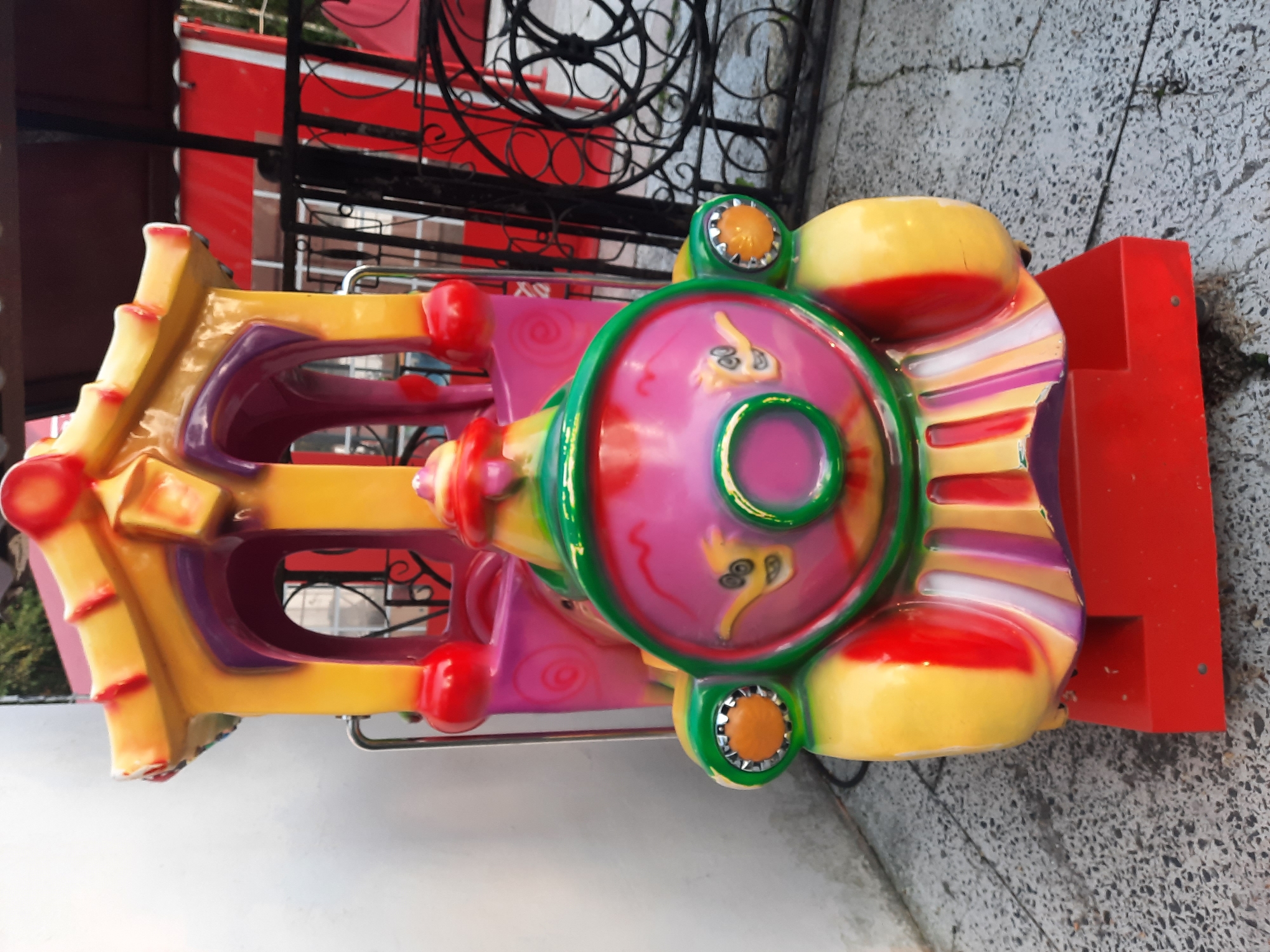 A brightly colored, vintage amusement park ride shaped like a train car is positioned outdoors on a grey, speckled paved surface. The ride is predominantly yellow, pink, and red, with purple accents and green trim. It features a whimsical design with painted cartoon eyes and a circular opening on the front. A red base supports the train car. In the background, a black wrought-iron fence separates the ride from red structures and greenery, suggesting an amusement park or carnival setting. The lighting indicates it is daytime, with shadows present. The location is specified as Unknown, Unknown.