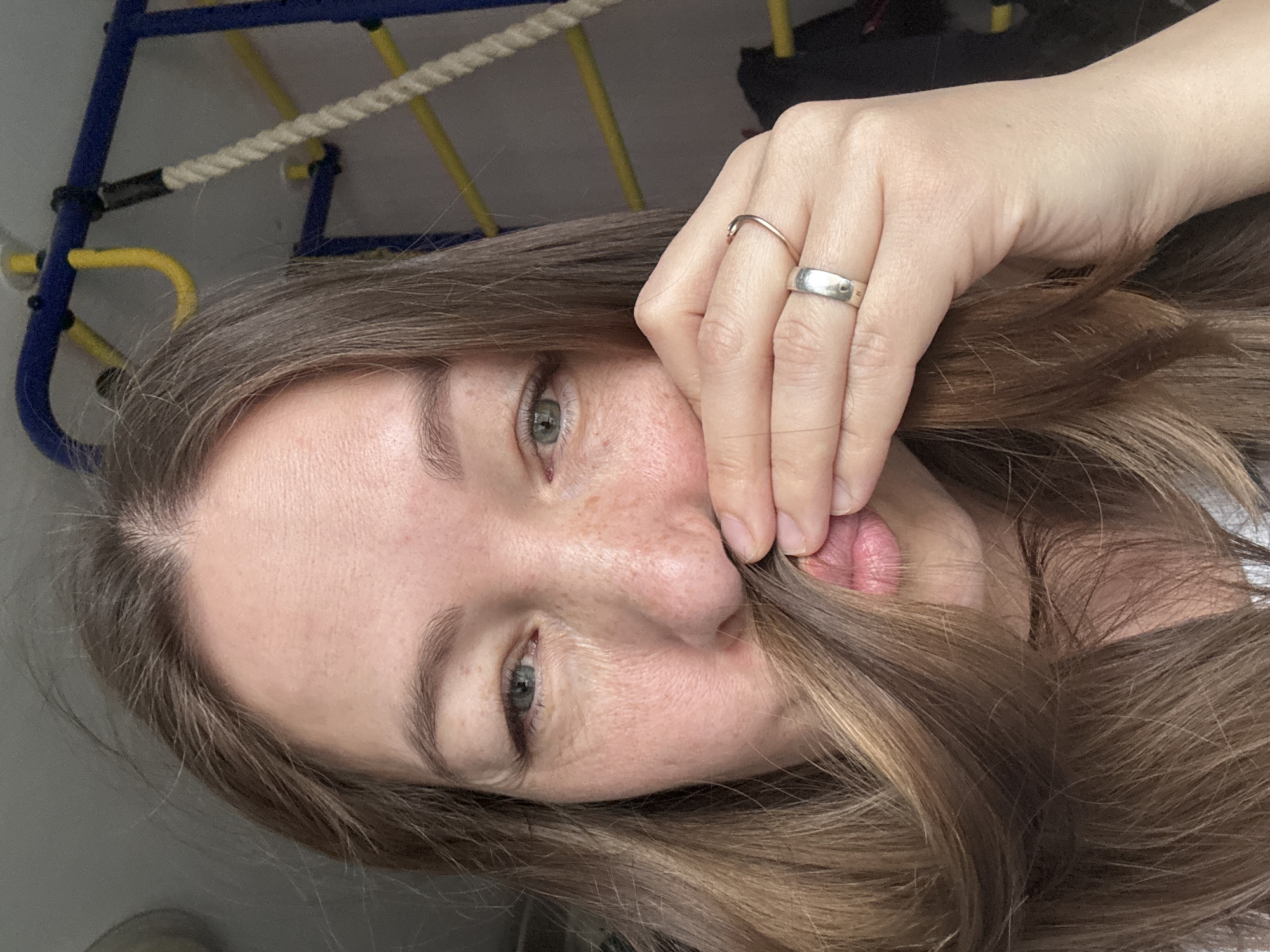 A woman's face is centered in the image, tilted slightly to the left, with her hand obscuring part of her mouth. She has light skin with freckles and light brown hair, part of which she's holding near her mouth. She has grey-green eyes. On her hand, there are two rings, a thin gold band and a wider silver band. In the background, a child's jungle gym structure with yellow and blue bars is visible. The location is possibly a private residence, but the exact location within the city of Chaikovsky, Russia, cannot be determined from the image. No specific actions or interactions are taking place in the image other than the woman posing.