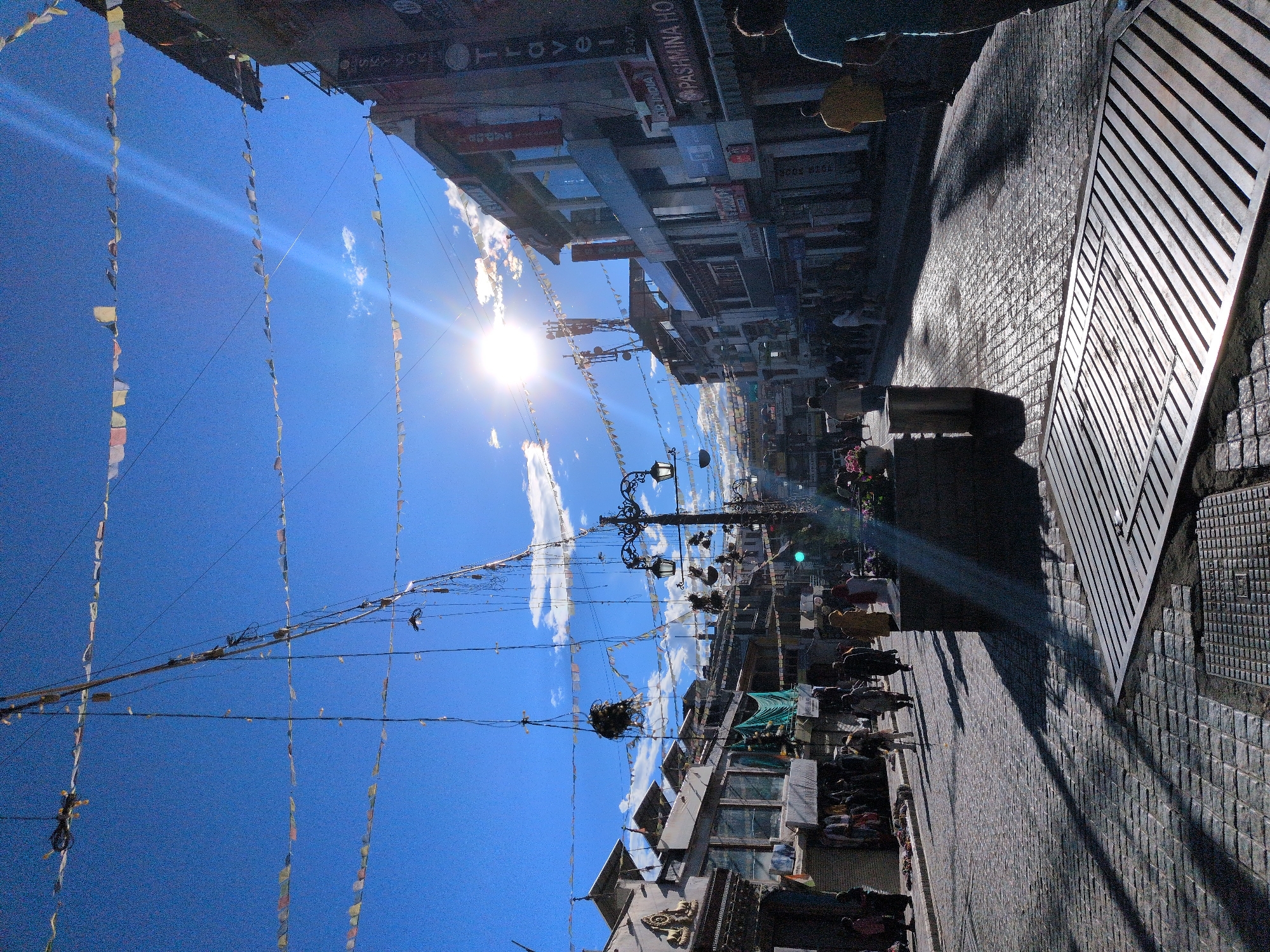A street scene in the city of Leh, India, is captured under a bright, sunny sky with scattered clouds. Colorful prayer flags are strung across the street, creating a festive atmosphere. The street is paved with cobblestones that reflect the sunlight, creating a glistening effect. Buildings line both sides of the street, many with multiple stories and awnings. People are walking along the street, some dressed in traditional attire. There are shops and stalls with goods displayed. The sun is high in the sky, casting long shadows. The overall impression is of a bustling marketplace in a mountainous region.
