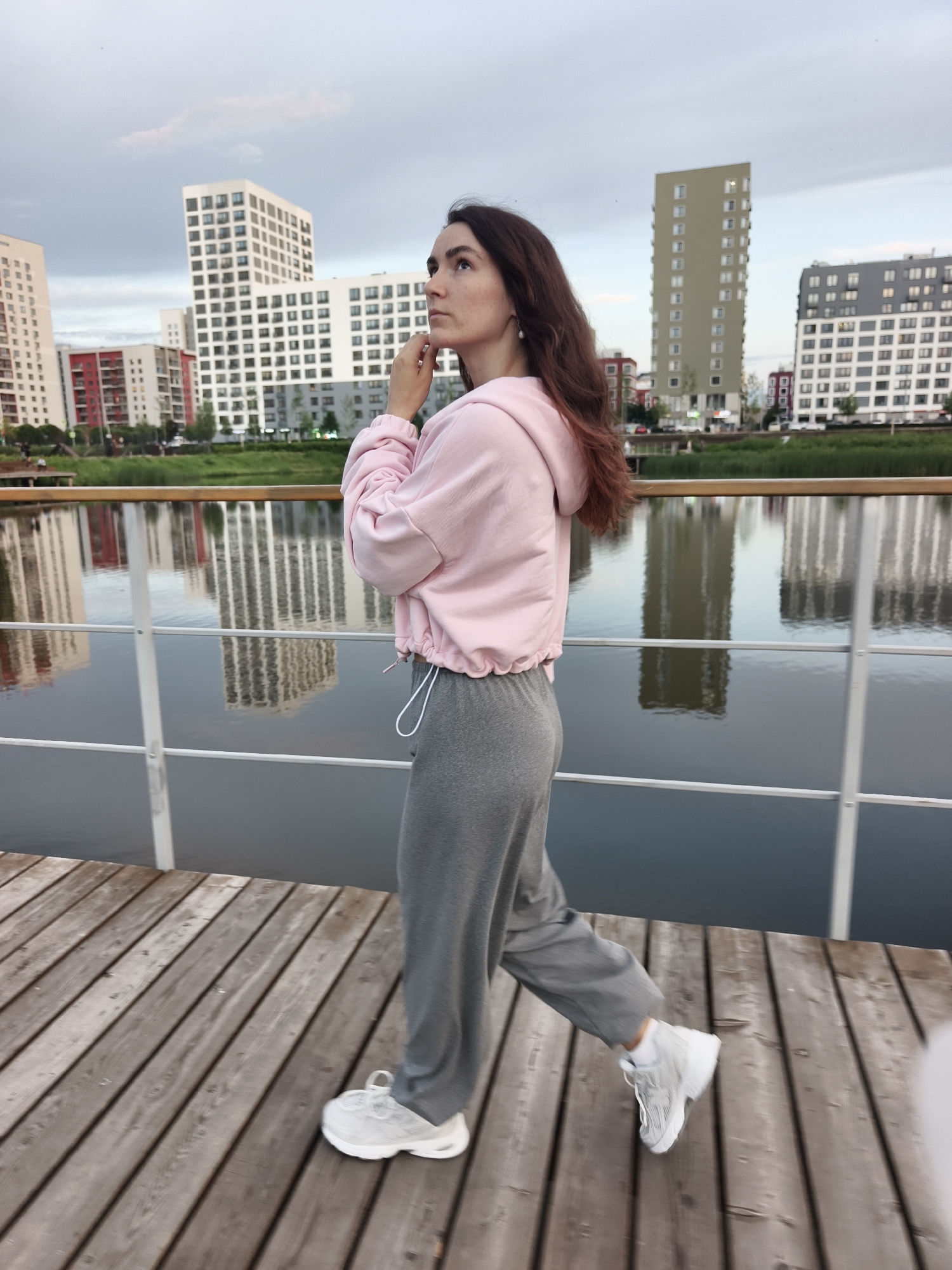 A woman with long, dark hair is walking on a wooden boardwalk next to a body of water. She is wearing a pink hooded sweatshirt and grey sweatpants, and white sneakers. In the background, there are several modern buildings with reflective windows, and the sky is cloudy. The scene appears to be in a city, possibly Tyumen, Russia, given the architectural style and the presence of a waterfront walkway. The overall atmosphere is calm and urban.