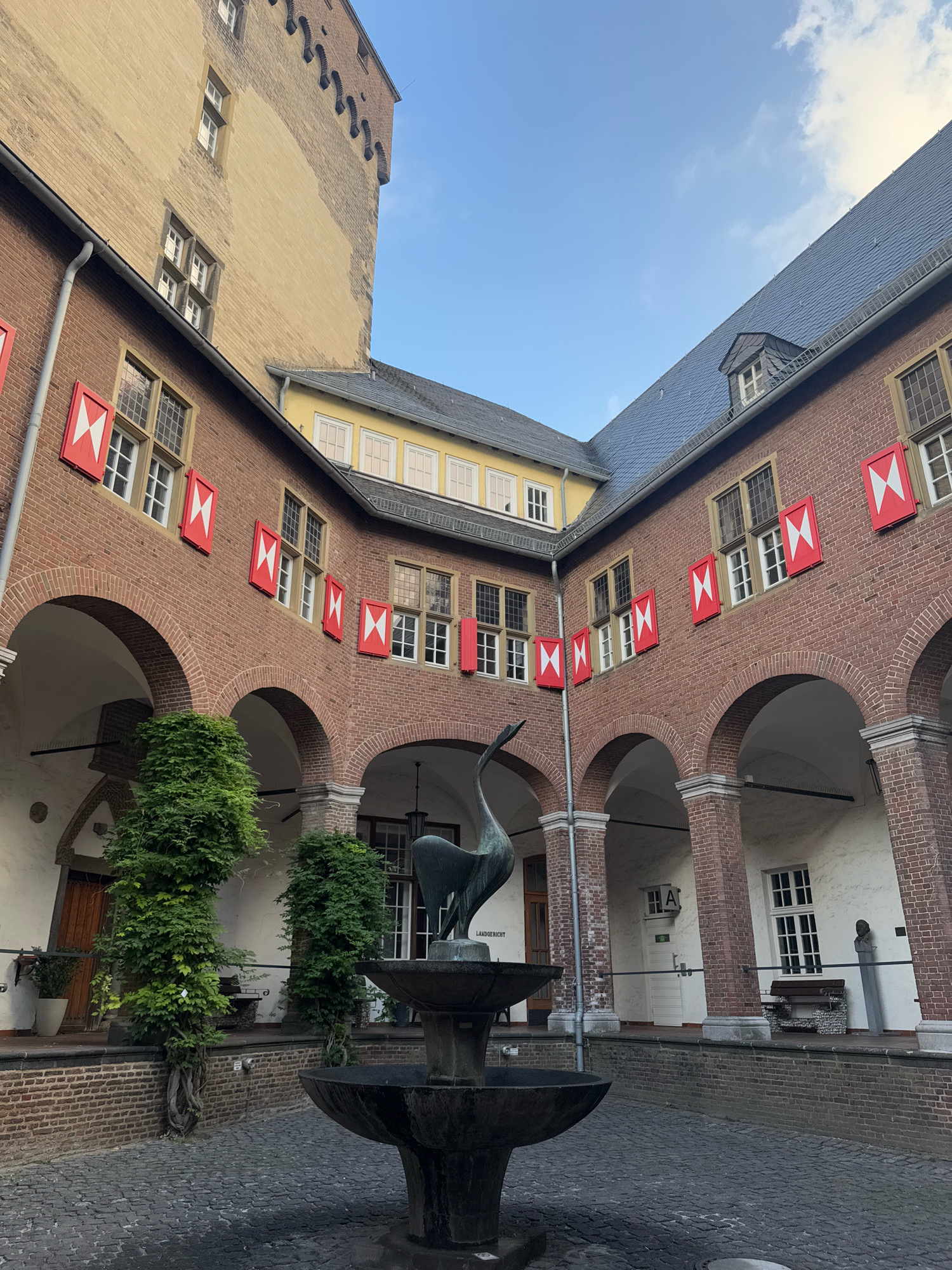 The image shows the inner courtyard of the Cleves Castle (Schloss Kleve) in Cleves, Germany. The courtyard is paved with cobblestones and features a multi-tiered fountain with a bird sculpture at its apex. The courtyard is surrounded by historic brick buildings with arched walkways. The buildings have distinctive red and white shutters on their windows, and some have visible gables and dormer windows. A tall, pale yellow tower with crenellations stands in the background on the left. Lush green ivy climbs some of the pillars in the arcade. A sign that reads "LANDGERICHT" is visible on a building to the right of the fountain. There are no people visible in the image. The sky above is a clear blue with a few scattered clouds.