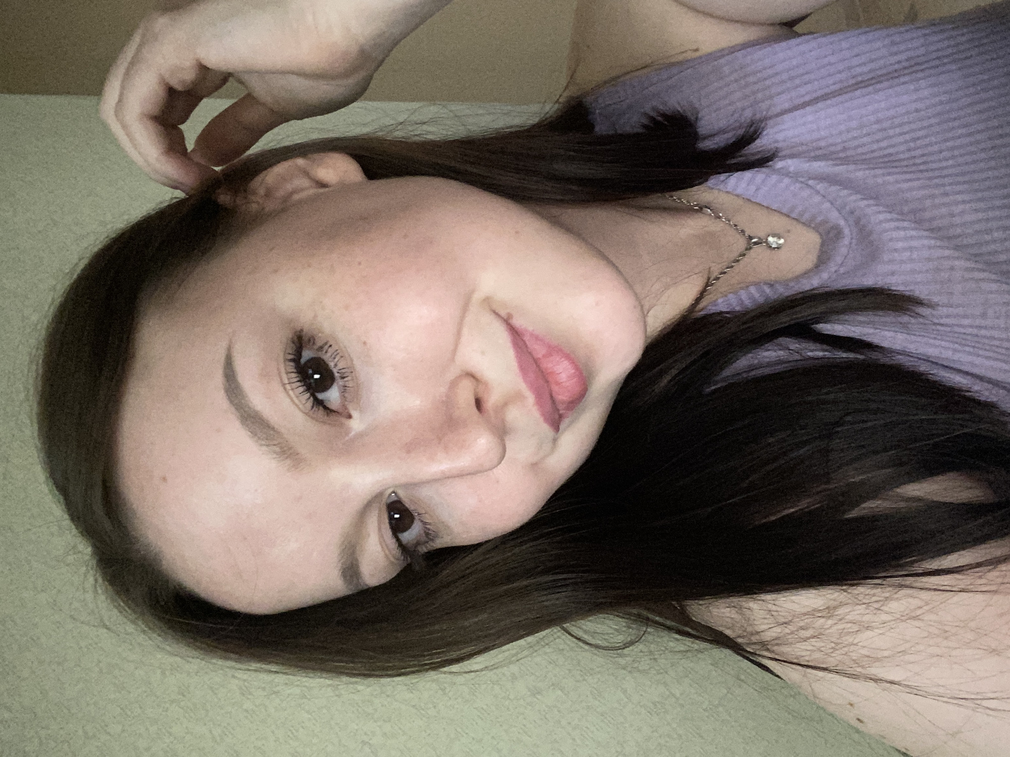 The image features a young woman lying down, looking directly at the viewer with her head tilted to the right. Her long, dark brown hair spreads around her head. She has fair skin, with freckles visible on her nose and cheeks. Her eyes are dark, and she wears mascara to accentuate her long lashes. Her lips are slightly parted, revealing a subtle pink color, and her expression is relaxed and gentle, with a hint of a soft smile.
Her right hand is raised, with her fingers lightly touching her hair near her ear. She is wearing a light purple, ribbed top, and a thin silver chain necklace with a small, clear pendant hangs around her neck. The background consists of a light, muted green or beige textured surface, suggesting an indoor setting like a bed or sofa. The lighting is soft, and no specific time of day or weather conditions are discernible. There is no visible text in the image. The scene conveys a calm and informal atmosphere, captured in Chaikovsky, Russia.