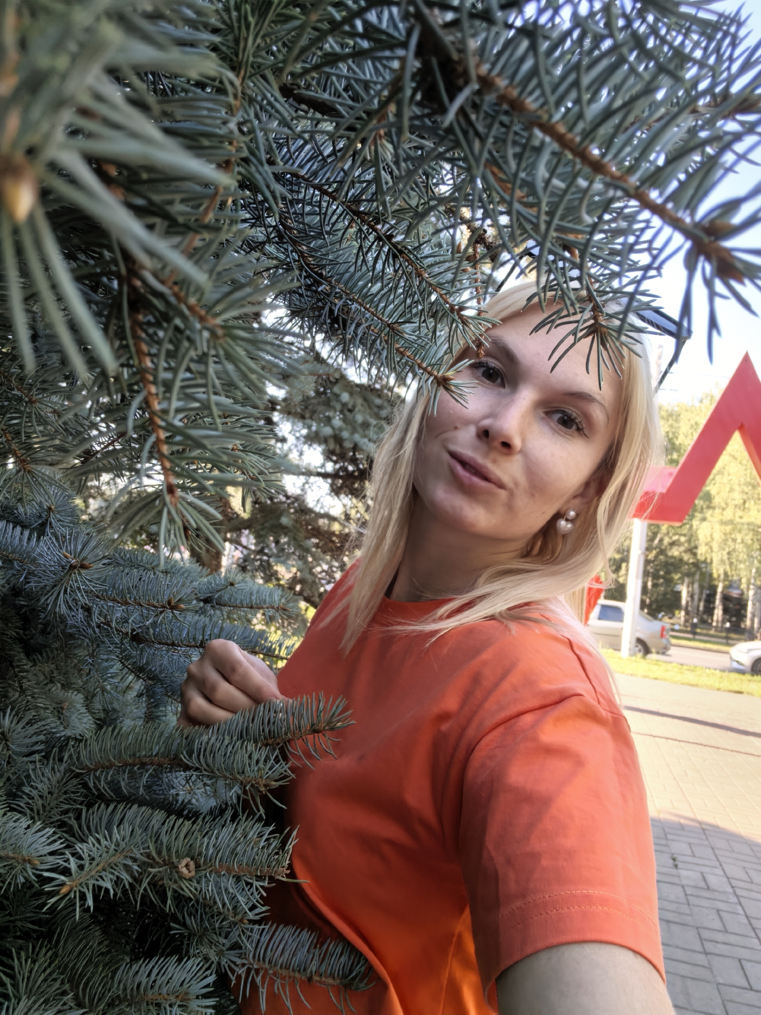 A young woman with blonde hair and fair skin is pictured from the chest up, partially obscured by the branches of a blue spruce tree. She is wearing an orange t-shirt and pearl earrings. The woman is looking towards the camera with a slight smile. In the background, there is a red triangular structure, trees, a sidewalk made of brick pavers, and a street with parked cars. The scene appears to be outdoors in an urban environment, possibly a park or a street in Sterlitamak, Russia, given the presence of the red triangular structure which is a common element of the Sterlitamak city sign. The lighting suggests it is daytime, with sunlight casting shadows.