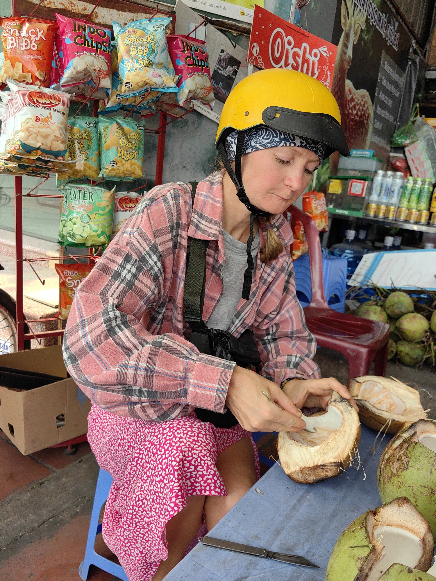 A woman wearing a yellow helmet and pink plaid shirt sits on a blue plastic stool and uses a small spoon to scoop the flesh from a halved coconut, one of several on a blue table. A knife lies nearby. Behind her are red and blue plastic chairs. The setting is a Vietnamese roadside food stand in Cao Xanh Ward, Vietnam. Stacks of coconut and various packaged snacks are displayed on the stand. Soft drinks and other items are also visible on the stand.