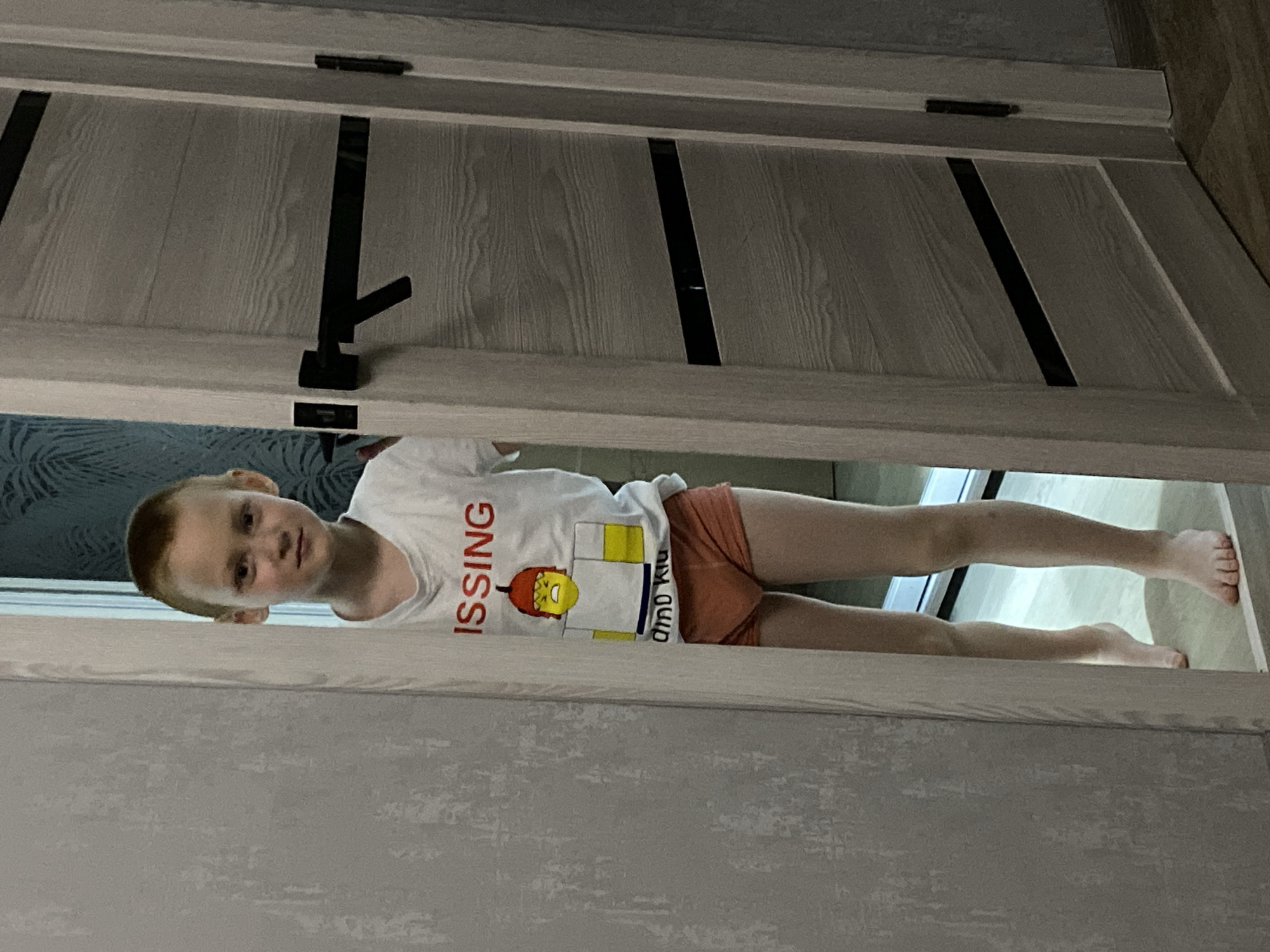 A young boy lies on the floor visible through the gap of a partially opened door. He is wearing a white t-shirt with a graphic of a Lego figure and the word "MISSING" on it, along with orange shorts. The door is grey with black design elements. The location is Noviy, Russia.