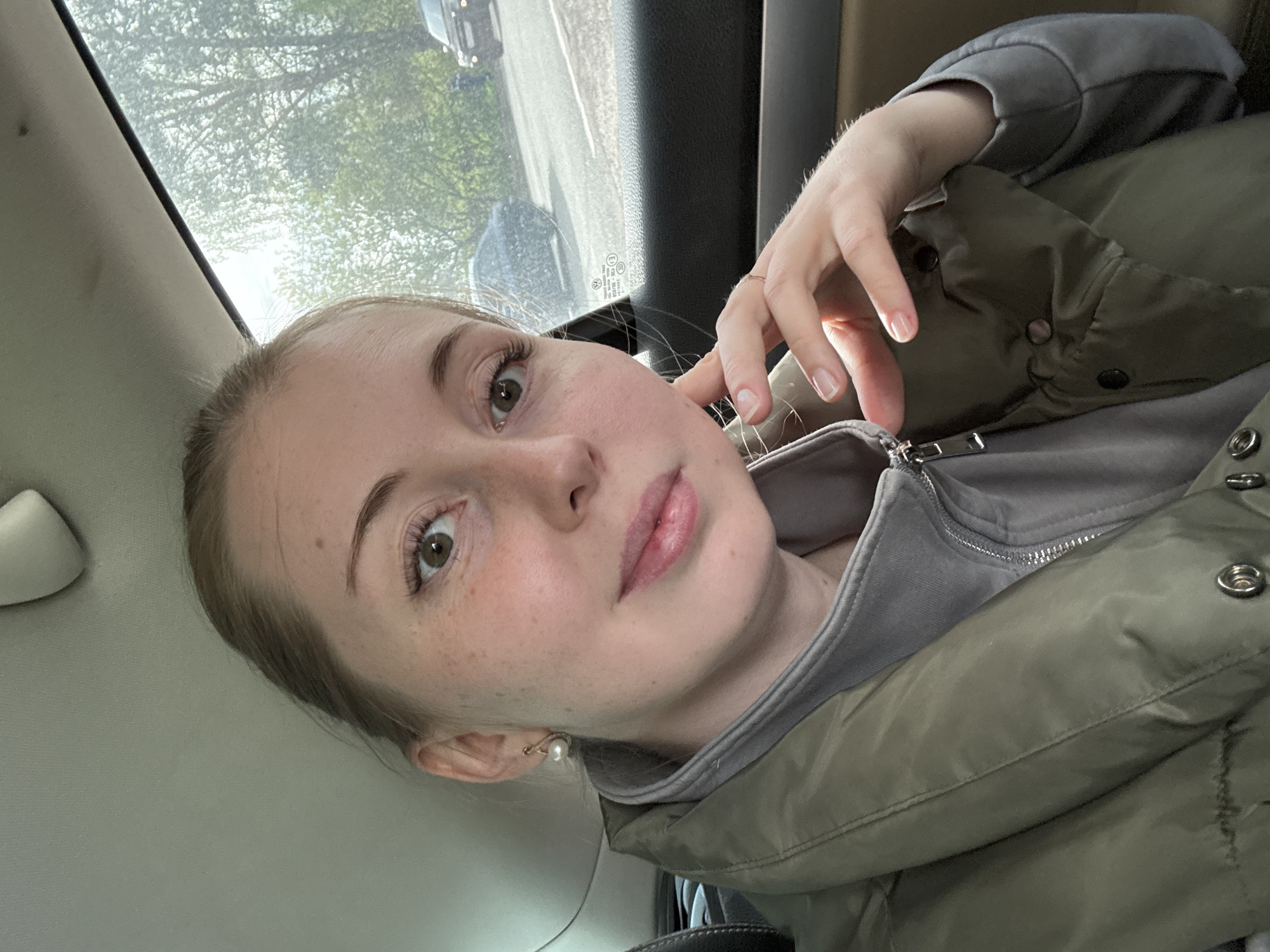 A fair-skinned young woman with freckles is in the passenger seat of a car. She has light brown hair pulled back, hazel eyes, and is wearing a pearl earring. Her hand, adorned with a simple gold ring, is near her face. She's wearing a gray zip-up hoodie under a green puffy jacket. The car window shows an indistinct view of trees and other vehicles. The location is Unknown, Unknown.