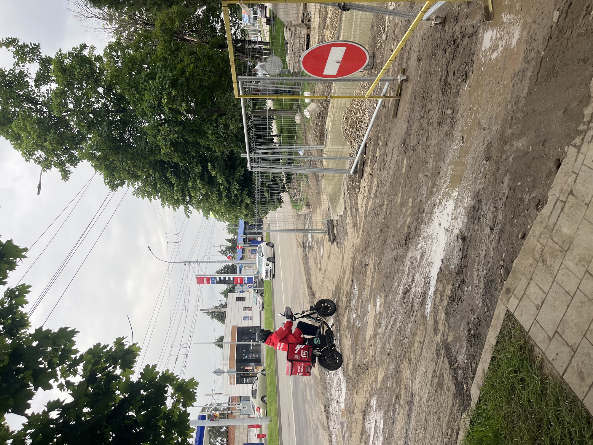 The image depicts a street scene in Krasnodar, Russia. A person wearing a red jacket and a helmet is riding a small electric scooter on a muddy, unfinished section of what appears to be a road or sidewalk under construction. The scooter has a delivery box attached. A red and white "no entry" sign is placed behind a metal fence, indicating a construction zone. In the background, there is a petrol station with "PETROL" signage, along with several buildings and passing vehicles. The sky is overcast, and trees line the street.