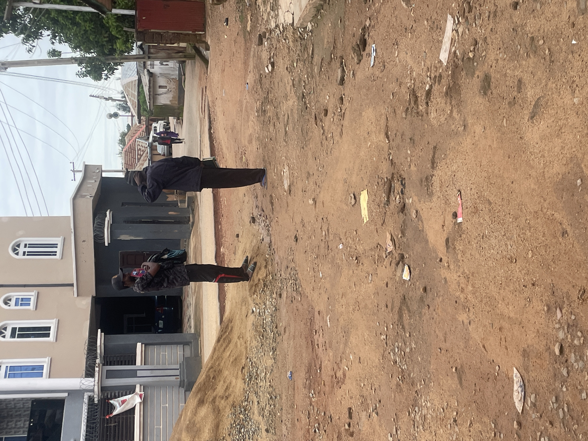 The image depicts a street scene in Jos, Nigeria. In the foreground, two men are standing on a dirt road. The man on the left is wearing a camouflage t-shirt, black pants, a black baseball cap, and is holding a backpack and a phone. The man on the right is wearing a dark jacket and black pants, with his back to the camera. The road is unpaved and littered with debris. In the background, there are buildings, some with corrugated iron roofs, and power lines. Trees and vegetation are visible, and the sky is overcast. The overall impression is of a dusty, somewhat neglected urban environment.