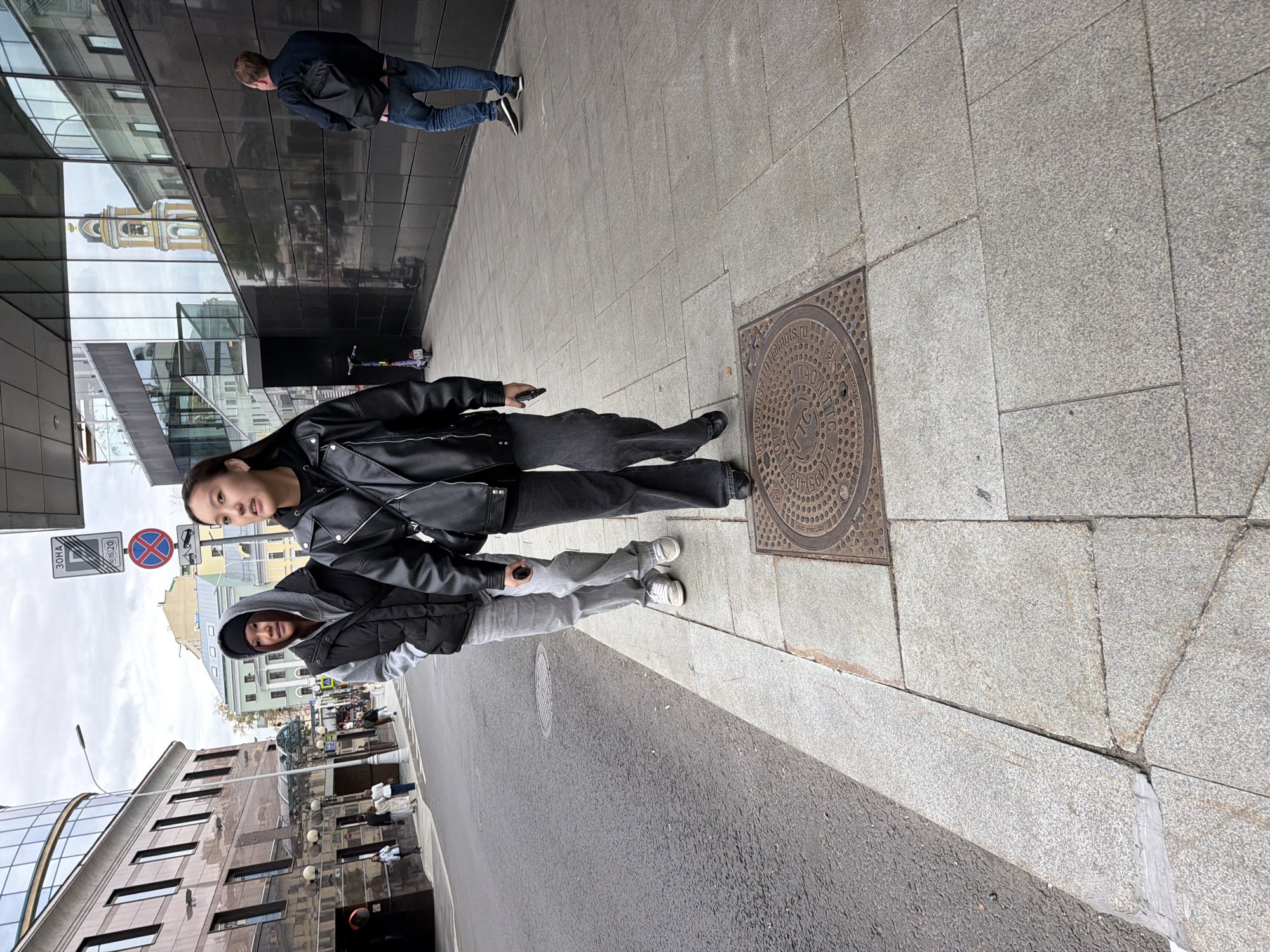 Two women walk on a sidewalk in front of a modern building with a glass facade. The woman on the left wears a grey hoodie, a black puffer vest, and grey jeans. The woman on the right wears a black leather jacket and dark grey jeans. A man walks further down the sidewalk behind them. The street is to the left of the sidewalk. The sky is overcast.