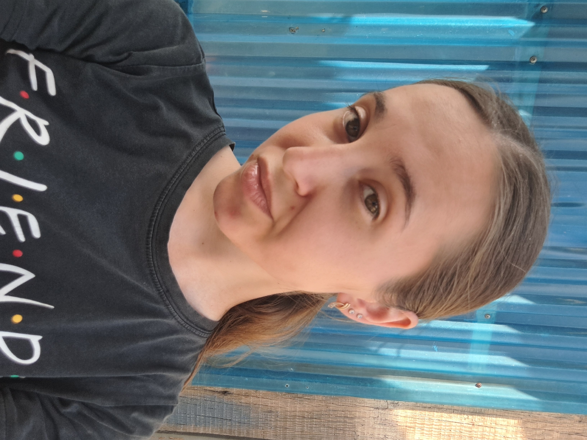 A woman with auburn hair pulled back in a ponytail is shown from the chest up, posing for a selfie. She is wearing a dark t-shirt with the word "FRIENDS" in white lettering, punctuated by colorful dots. The woman has light skin and light brown eyes, and is wearing a small gold earring with a star shape and a small diamond in her left ear. She is standing in front of a corrugated blue metal wall, with a section of weathered wooden siding visible to the right of the wall. The scene appears to be outdoors. The woman is located in Vypasne, Ukraine.