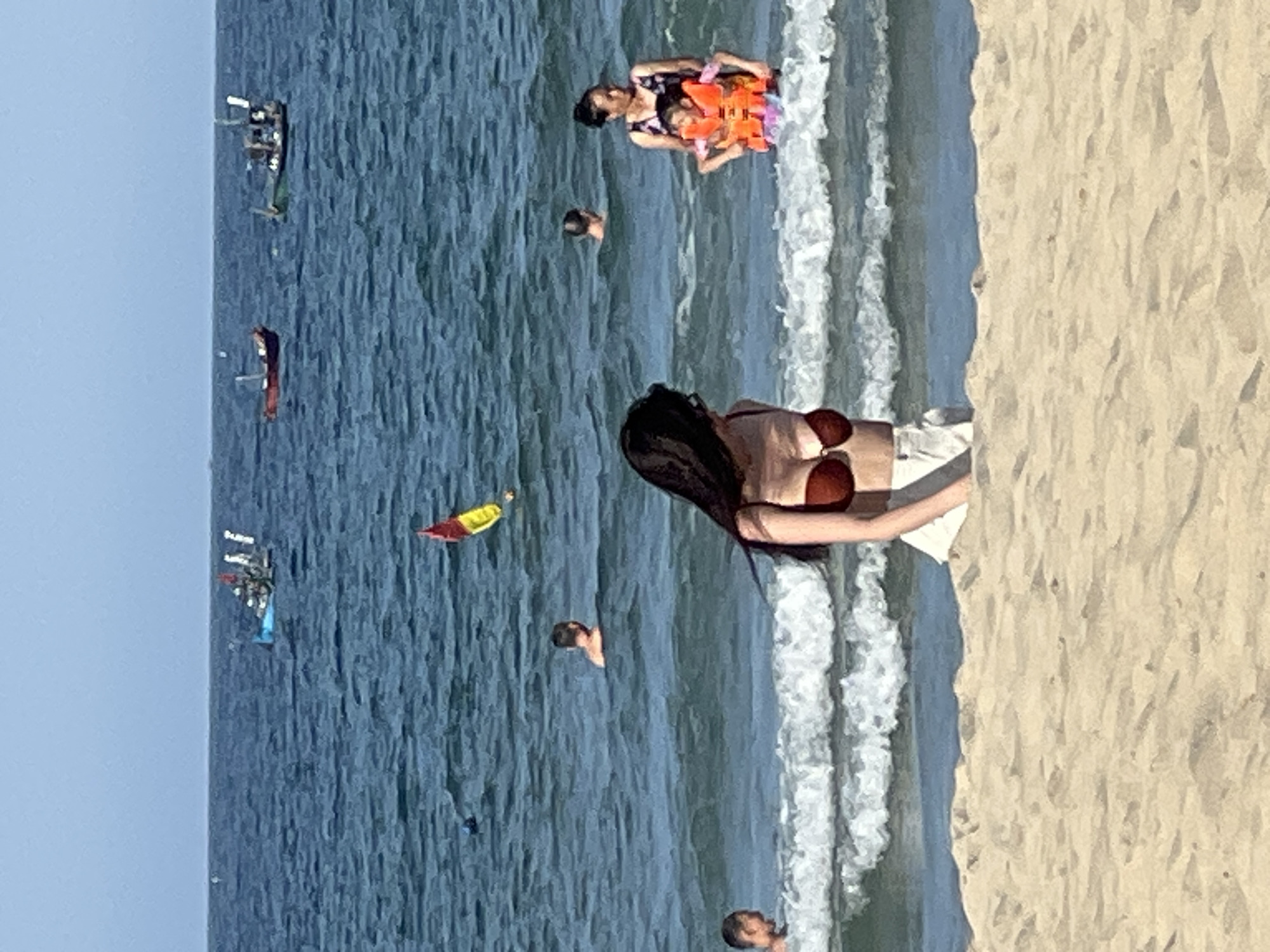 The image depicts a sunny day at a beach in Đà Nẵng, Vietnam. The scene is divided into the sandy shore on the right and the ocean on the left. Several people are visible, enjoying the water and the beach.
In the water, a woman in a bikini is standing near the shore, facing away from the camera. She appears to be looking out at the ocean. Further out in the water, a woman and a child are swimming. The child is wearing an orange life vest. Other individuals can be seen swimming or wading in the water, with only their heads or upper bodies visible. There are also several small boats or jet skis in the distance, indicating recreational water activities. A red and yellow flag is flying in the water.
The sand on the beach is light brown and appears to be rippled by the wind or water. The ocean is a deep blue, with white waves crashing along the shore. The sky is clear and light blue, suggesting fair weather. The overall mood of the image is relaxed and leisurely, capturing a typical beach scene on a warm day. No visible text is present in the image.