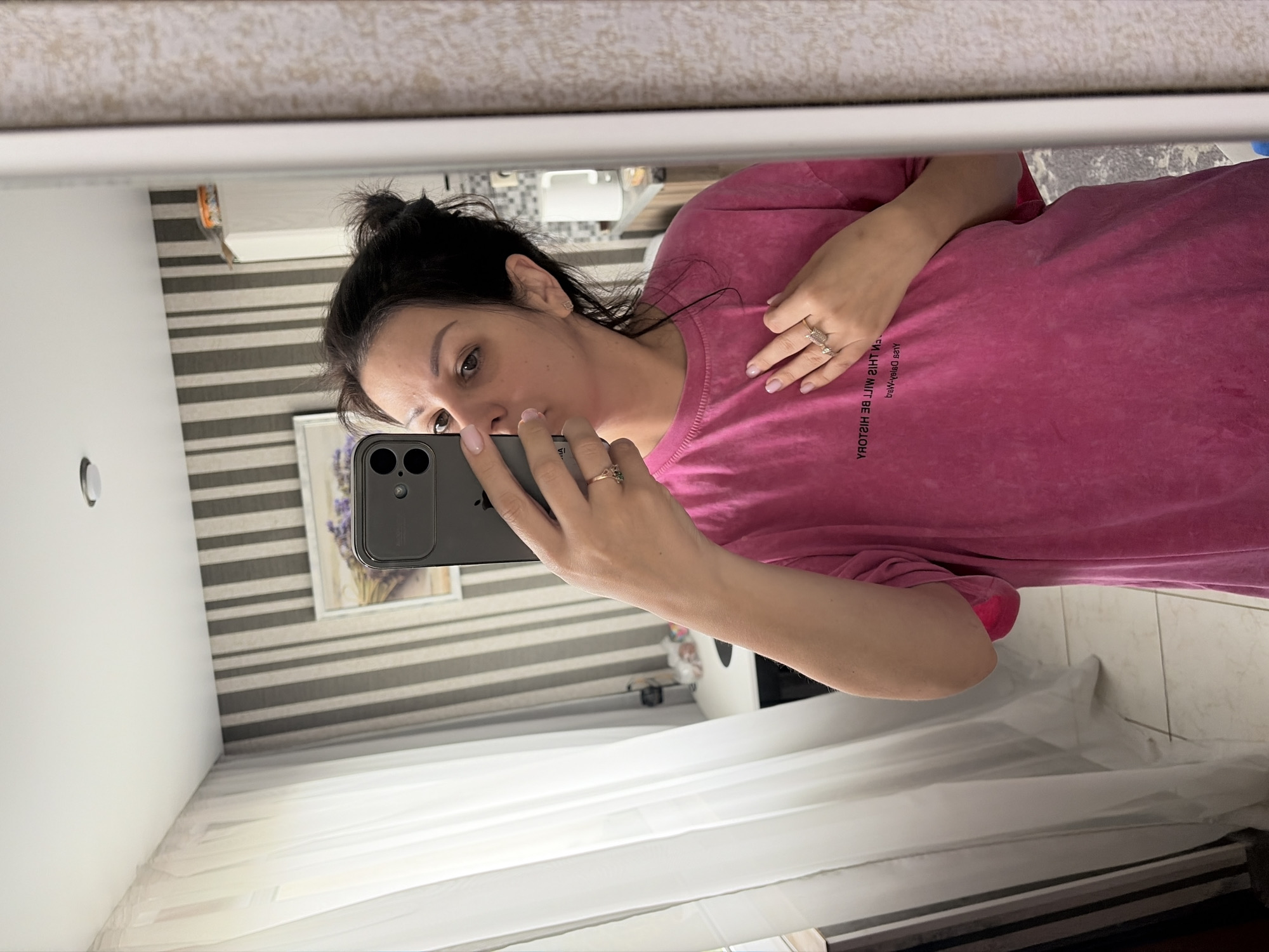 A woman with dark hair pulled into a bun is taking a mirror selfie in a room that appears to be in Sochi, Russia. She is wearing a bright pink t-shirt with text on it. Her left hand is holding her phone, and her right hand is on her chest. The background shows striped wallpaper, a window with sheer white curtains, and a white cabinet. There is a framed picture on the wall and some items on top of the cabinet.
