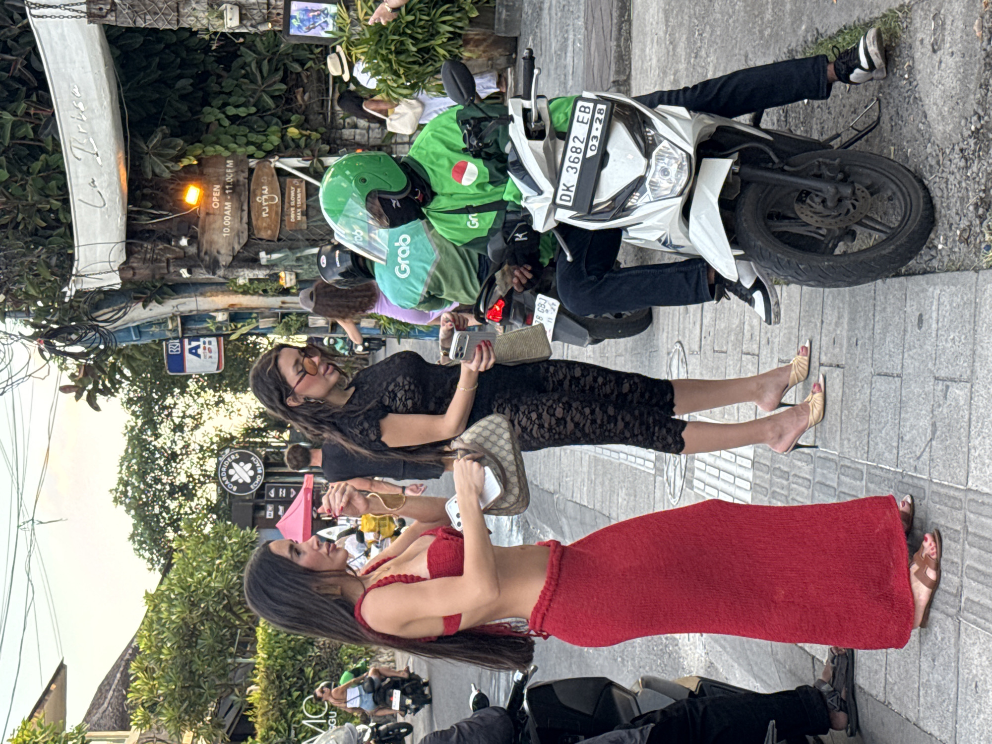 The image captures a street scene in North Kuta, Indonesia. Two women are standing on the sidewalk next to parked motorbikes. One woman is wearing a red dress and the other a black lace dress. They are facing each other and appear to be in conversation. One is holding a phone and the other is holding a small handbag. Next to them, two motorbikes are parked. One is ridden by a man in a green uniform with "Grab" written on the back. The license plate reads "DK 3682 EB". In the background, there's a sign for "La Brisa" with the opening hours "10.00AM-11.00FM". Other signs and businesses are visible in the background.