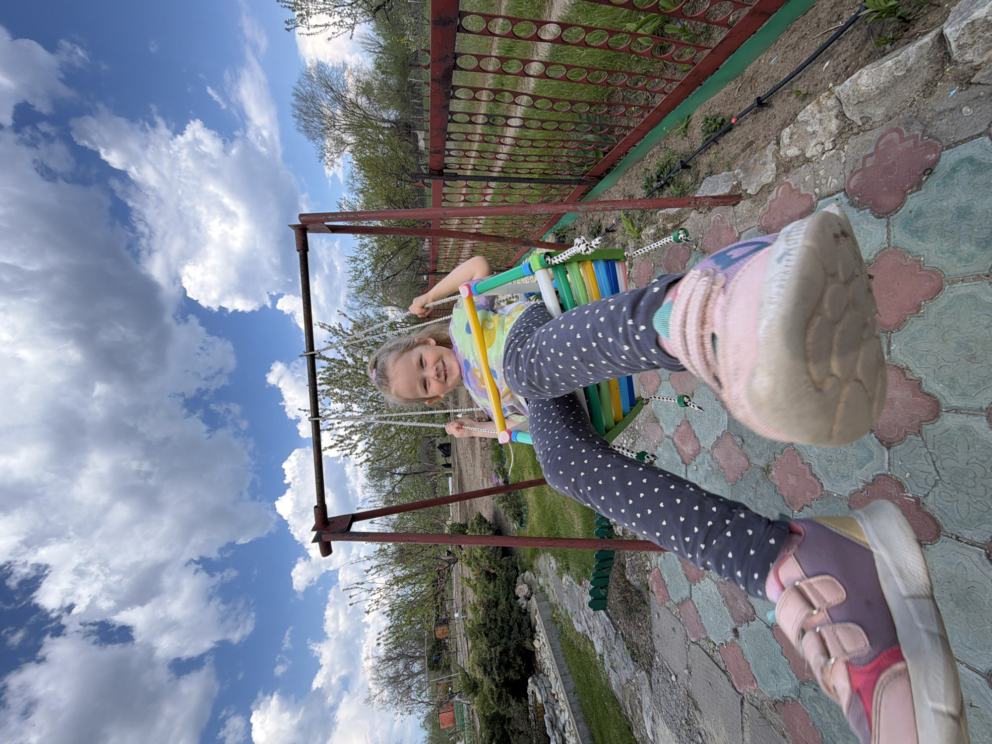 A young girl is captured mid-swing, her body angled towards the camera. She has blond hair and is wearing a multi-colored top and dark leggings with white polka dots. She wears a shoe on each foot. The swing is made of colorful planks. The swing set appears to be in a backyard setting in Kamyshin, Russia, with a metal fence and greenery visible in the background. The sky above is partly cloudy. The ground beneath the swing is paved with decorative tiles.