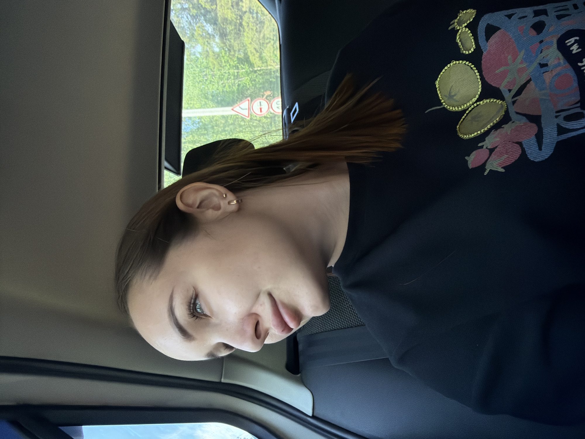 A young woman is in a car. She has light skin, blue eyes, and brown hair. She is wearing a dark, long-sleeved shirt with colorful floral and text designs. The car is traveling on a road in the city of Chaikovsky, Russia. A road sign is visible through the window. It indicates a speed limit of 40 km/h and a turn. The surrounding environment appears to be green with trees.