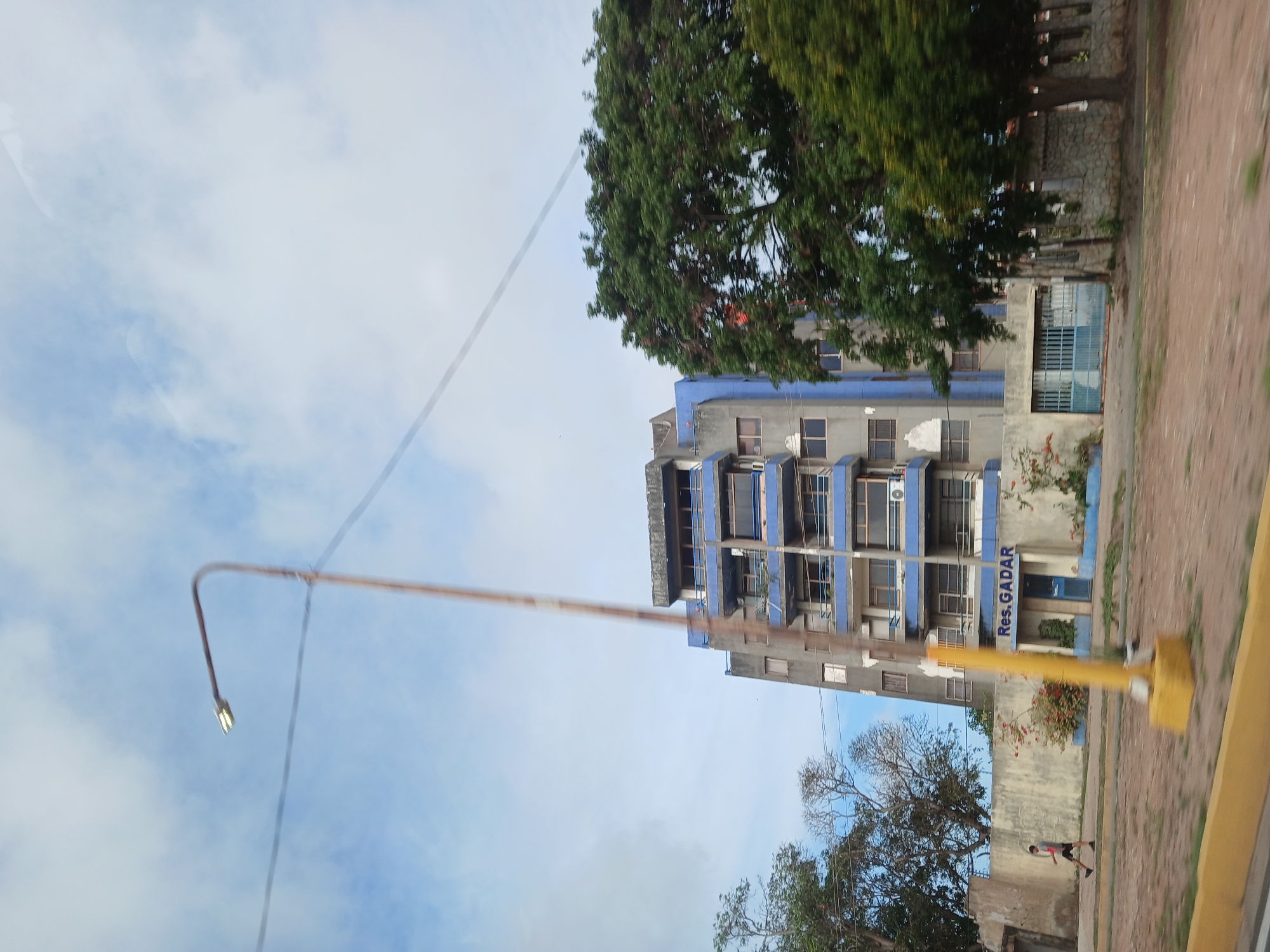 An eye-level, wide shot captures a multi-story apartment building in El Valle del Espíritu Santo, Venezuela. The building, predominantly gray with blue trim, features several balconies with blue railings. The words "Res. GADAR" are visible in blue lettering above the entrance on the ground floor. A person in dark shorts and a gray shirt is walking on a paved path in front of the building. A tall, yellow streetlamp with a light fixture stands prominently in the foreground. Adjacent to the building, a large, leafy green tree dominates the right side of the frame. The sky above is a mix of blue and scattered white clouds. The overall scene suggests an urban environment with residential structures and natural elements.