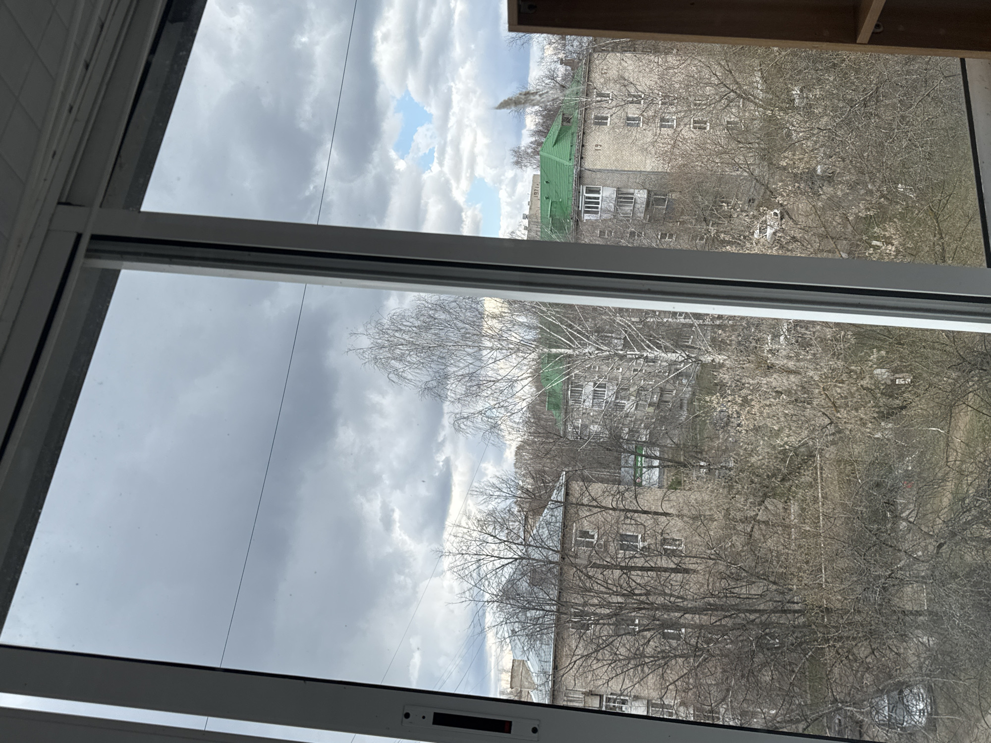 The image is a view from inside a building, looking out through a window towards the city of Chaikovsky, Russia. The scene is dominated by gray, overcast skies with glimpses of blue sky breaking through the clouds. Buildings, characterized by their Soviet-era architecture, are visible through the bare branches of trees. The buildings are generally multi-story structures, and one has a distinctive green roof. The environment suggests a late winter or early spring season due to the lack of foliage on the trees. The details of the city are partially obscured by the branches of the trees in the foreground. No people are visible in the frame. The focus is on the urban landscape seen from a window in Chaikovsky under cloudy skies.