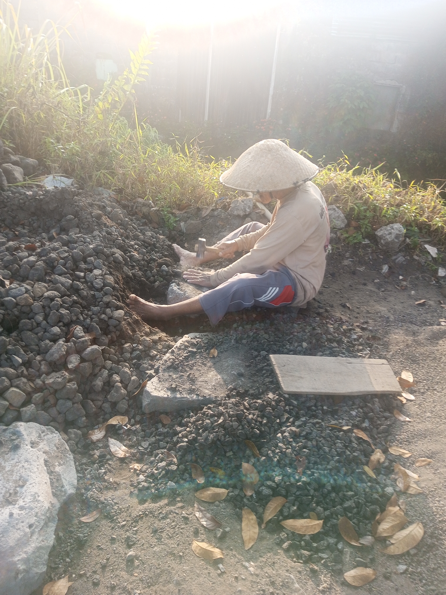 In Garung, Indonesia, a person wearing a conical straw hat and a light-colored long-sleeved shirt and grey pants with red and white stripes sits on the ground amidst a pile of grey gravel. Their right hand is holding a hammer, and their left hand is positioned near their lap, possibly resting. Their bare feet are visible, with the right foot digging into the gravel. A wooden plank lies flat on the ground in front of them. The environment is outdoors, with dry vegetation and scattered brown leaves visible in the background and foreground. The scene is illuminated by bright, hazy sunlight, suggesting it is either early morning or late afternoon.