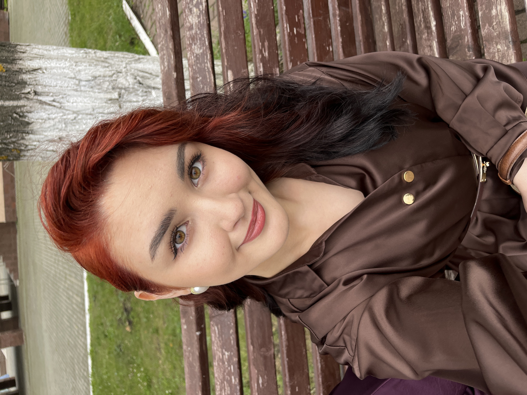 A woman with auburn and black hair is sitting on a wooden bench outdoors. She is wearing a brown satin top with gold buttons. The woman is looking at the camera and smiling. Her eye makeup is visible. She is wearing a pearl earring. The background shows a tree trunk and green grass. The photo appears to have been taken during the day, with natural lighting. The overall mood is casual and relaxed.
