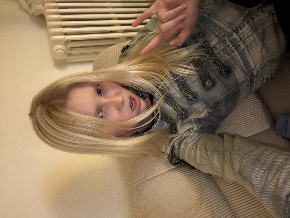 A young adult female with long, straight blonde hair and a light complexion is depicted in a close-up, selfie-style photograph, seated indoors on a light-colored, ribbed sofa. She wears a light-wash denim jacket, styled with two vertical rows of decorative braiding and metallic buttons down the front, and a metallic chain detail on the left shoulder. Her facial expression is intense, characterized by narrowed blue eyes and an open mouth revealing teeth. Her left hand is raised, performing the "sign of the horns" gesture. The background features a plain light-colored wall on the left and a white cast-iron radiator on the right, indicating an interior room within the city of Paris, France. A dark blue object is partially visible on the sofa in the lower center.
