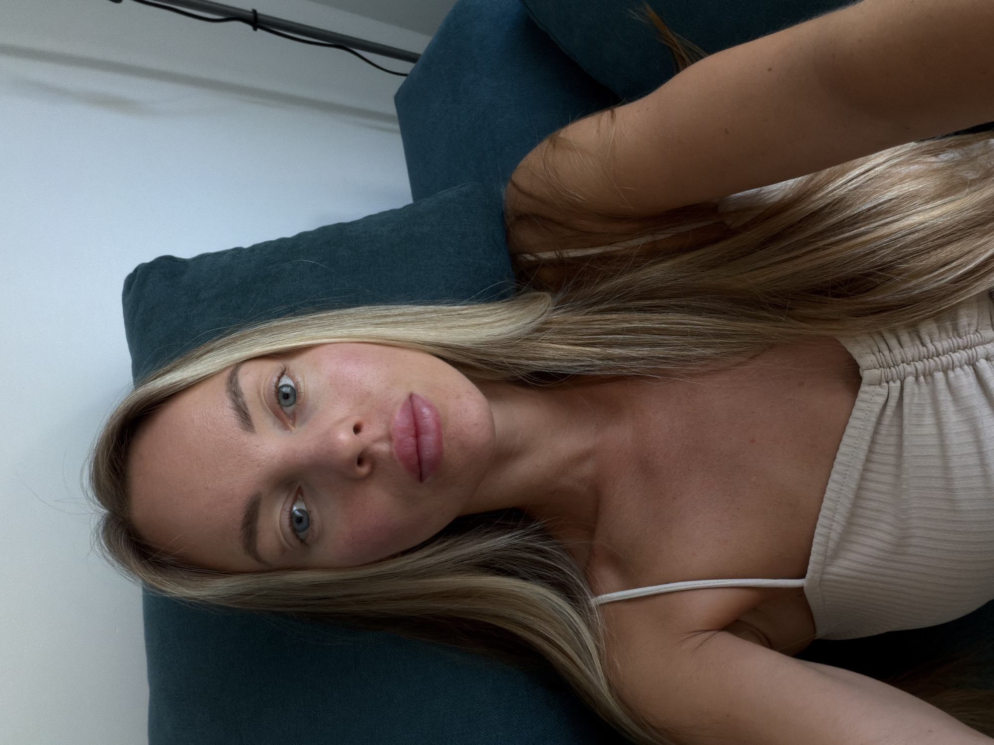 An indoor, waist-up, selfie-style portrait shows a woman with long, blonde hair, blue eyes, and light skin. She is wearing a beige, ribbed tank top with thin straps and a ruched neckline. The woman is seated on a dark teal couch, with the back of the couch visible behind her. The wall behind the couch is light-colored. A black lamp cord is visible on the right side of the frame. The lighting is soft and natural, suggesting it is daytime. The location is identified as Finestrat, Spain.