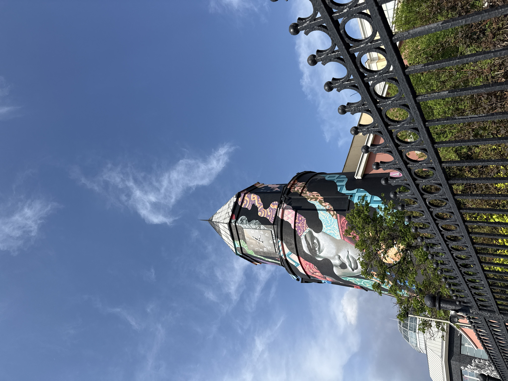 The image shows a view of the clock tower of the puppet theater named after Sergey Obraztsov, located in Moscow, Russia. The tower is painted with colorful murals, including a large face. In the foreground, there is a black wrought-iron fence. The sky is blue with light clouds.