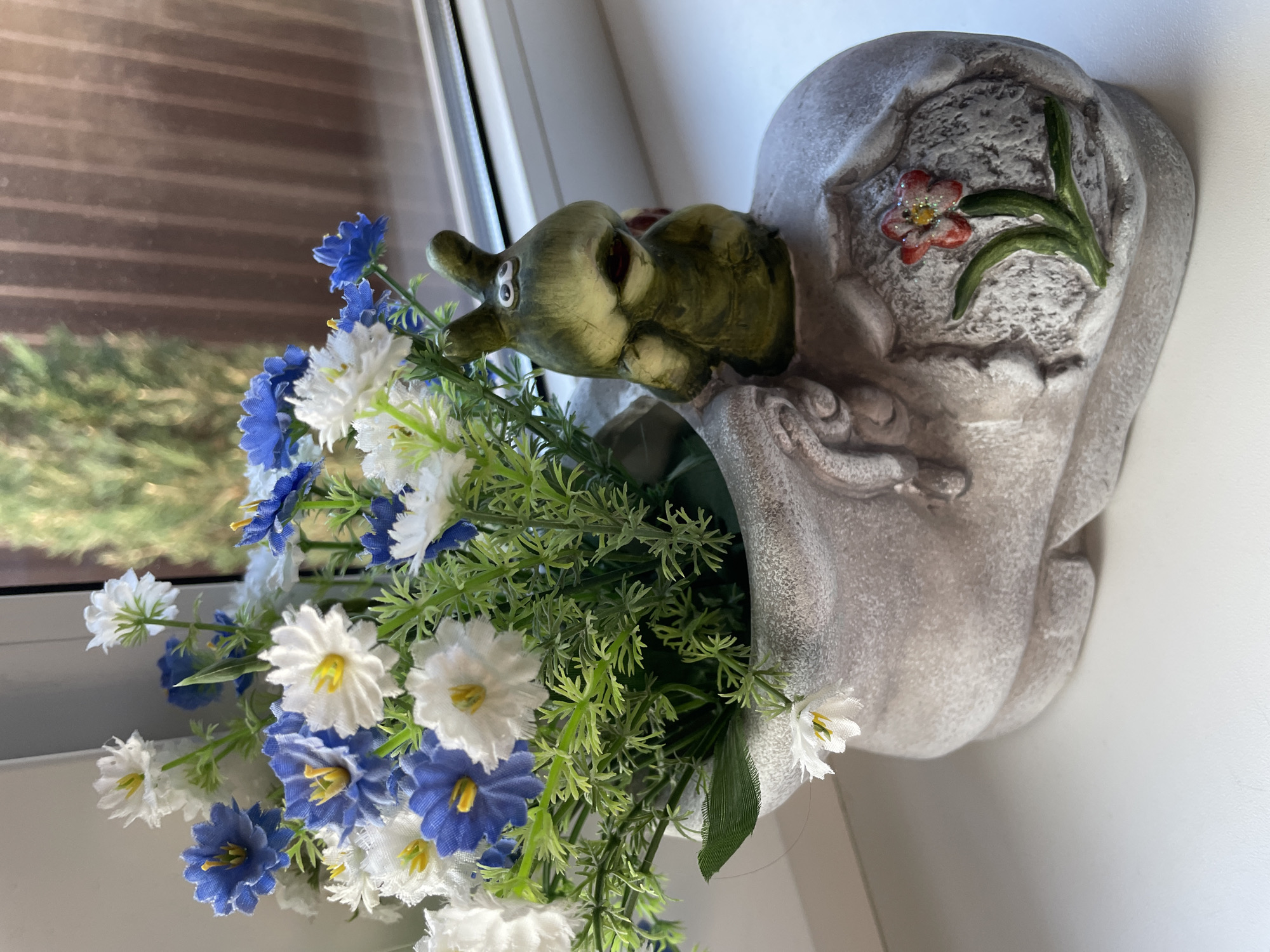 Here is a detailed description of the image:
The image shows a decorative flower arrangement in a pot shaped like a frog. The pot is grayish in color and appears to be made of stone or ceramic, with a whimsical design incorporating a frog's head and body. The frog's head is green with darker accents.
Arranged in the pot are artificial flowers in shades of white and blue, interspersed with green foliage. The arrangement is full and overflowing from the pot. The scene is lit by natural light.
The image was taken indoors, likely on a windowsill or a surface near a window, as suggested by the window frame and light filtering through. The location is identified as Лесной Пейзаж Кп (шарапово) in Russia.