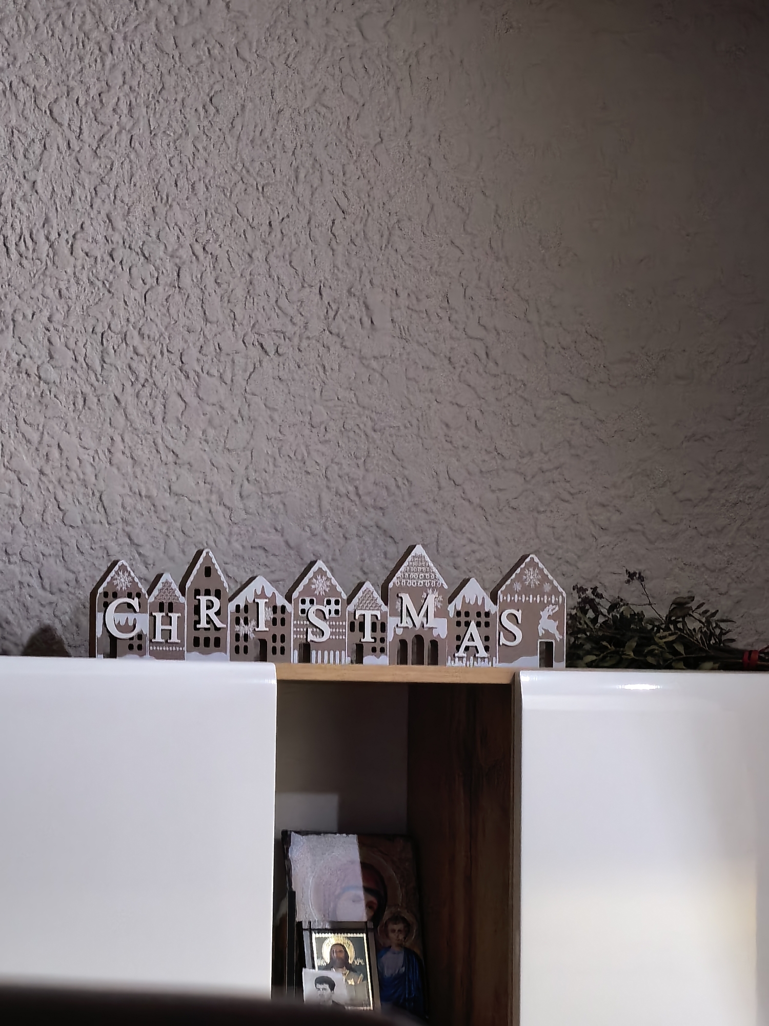 A medium-shot photograph shows a "CHRISTMAS" decoration on a shelf above white cabinets. The letters of the word are displayed on miniature gingerbread house-shaped pieces, which are brown with white trim and text. To the right of the gingerbread houses is a bundle of leafy plant branches. Behind the shelf is a gray wall with a textured surface. Under the shelf and inside the cabinet are two framed pictures. The cabinets have a white, glossy finish and take up the bottom part of the image. The room is dimly lit.