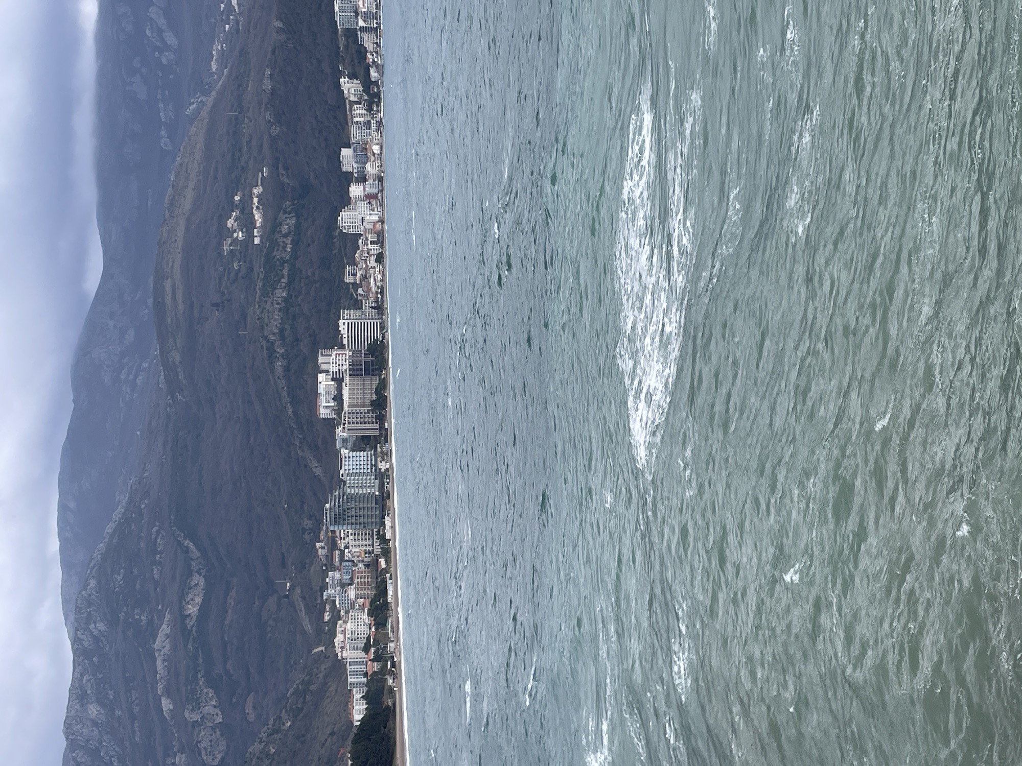 The image presents a panoramic view of the city of Budva, Montenegro, observed from the sea. The lower half of the frame is occupied by a body of choppy, dark teal-colored water, exhibiting numerous small waves and scattered patches of white foam on its surface. Along the midground, a sandy beach forms the coastline, backed by a dense urban development. This cityscape comprises a horizontal expanse of multi-story buildings, including modern white and light-colored high-rises, residential blocks, and other commercial structures. In the background, a range of steep, rugged mountains rises prominently. These mountains are predominantly covered in dark, sparse vegetation, and their upper sections are obscured by low-hanging clouds or mist, indicating an overcast sky. Small clusters of buildings are discernible on the lower slopes of the mountains. No individuals or specific activities are visible within the frame.