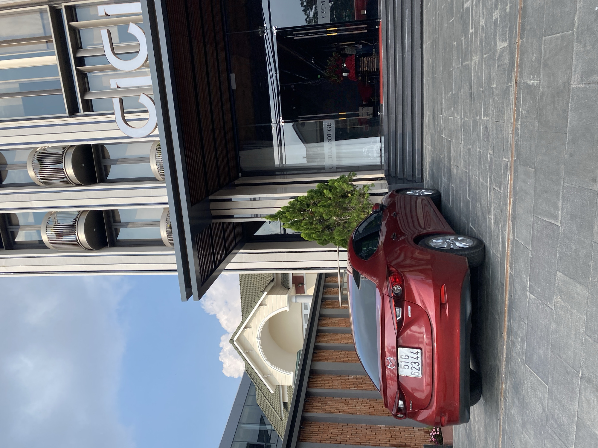 The image depicts a red Mazda parked on a grey stone-paved area in front of a building. The building features modern architecture with vertical metal strips and circular vents. The name "UDIC" is visible on the building's facade. The entrance to the building is marked by glass doors with the phrase "O'CHAU ROUGE" visible on one of the doors. A small green tree is positioned near the entrance. Part of a house with a white arched entryway and green roof is visible in the background. The license plate on the car reads "51G 62344". Based on the architectural style and signage, the location is likely in the city of Da Lat, Vietnam.
