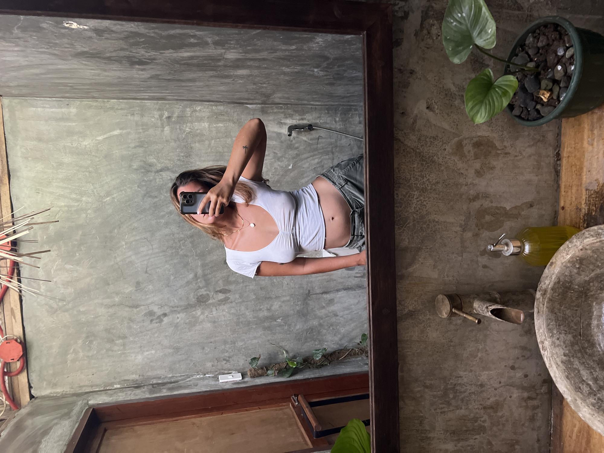 A woman, visible in a mirror reflection, takes a selfie in a bathroom. She is wearing a white, cropped V-neck top and grey pants. Her hair is light brown and falls around her shoulders. She has a necklace with a pearl pendant. The bathroom walls are made of a rough, grey concrete material. To the left of the mirror, a wooden door is visible, and a green vine climbs a moss-covered pole. Below the mirror, a stone sink with a brass faucet is visible, along with a pump bottle of soap and a potted plant with two large green leaves. The scene appears to be in General Luna, Philippines, as indicated by the context provided.