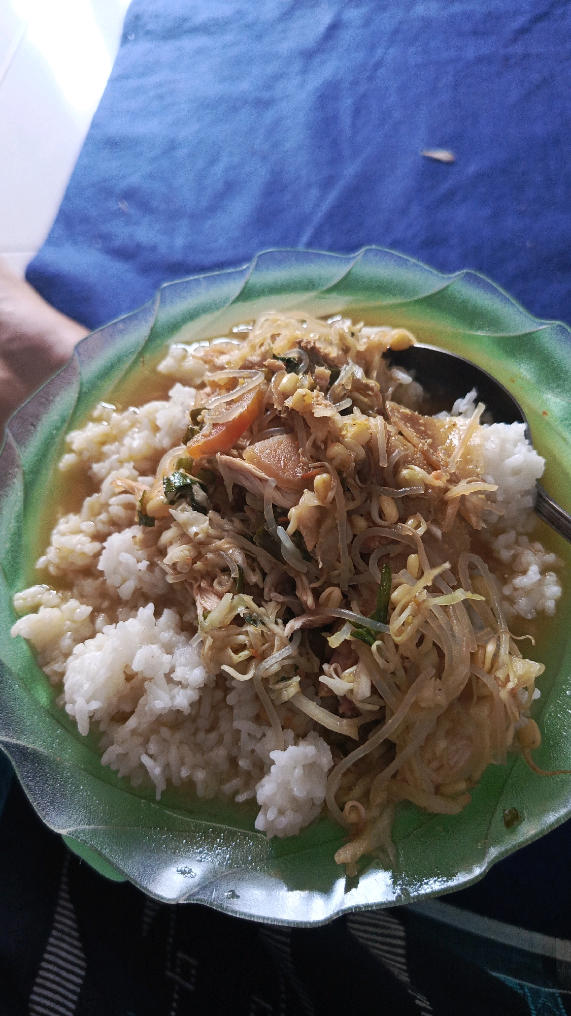 In Babadan, Indonesia, a green, translucent, wavy-edged plate holds a serving of rice topped with shredded chicken and bean sprouts in a savory broth. A spoon rests on the right side of the plate, submerged in the broth. The background consists of a blurred blue textile and a sliver of a white surface.
