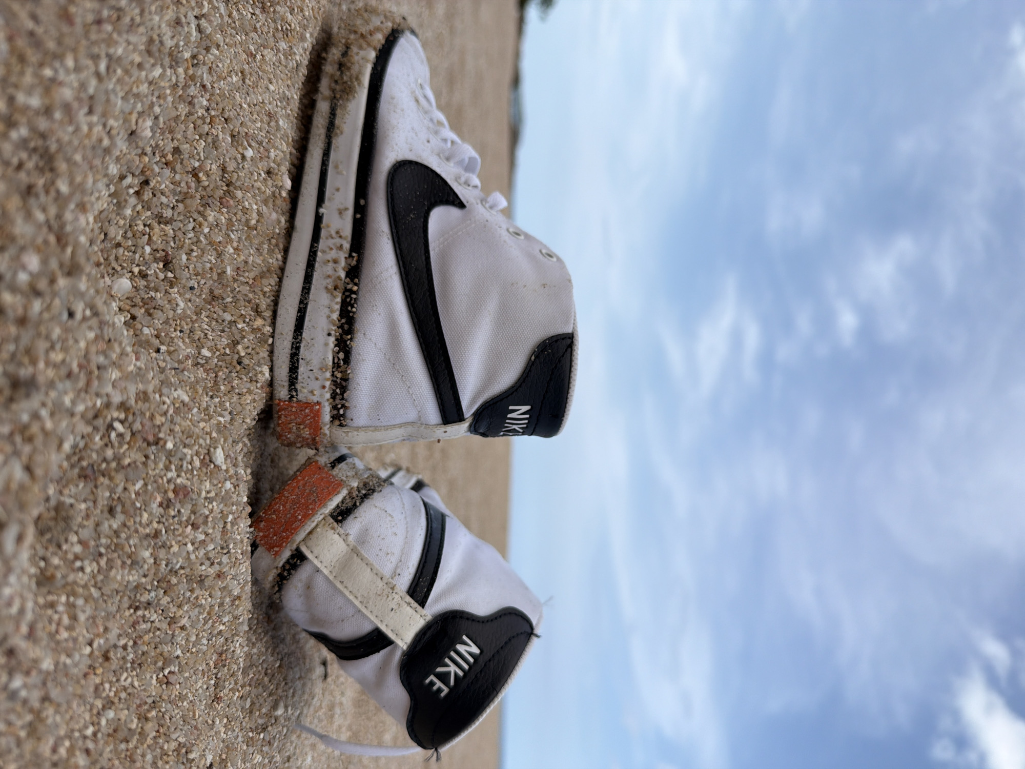 A pair of white Nike high-top sneakers, partially covered in sand, are placed on a sandy beach with the ocean and sky in the background. The sneakers are positioned at an angle, with the left shoe closer to the foreground and facing left, and the right shoe angled away from the viewer to the right. Both shoes display the Nike logo. The beach is composed of fine sand and small pebbles. The sky is a light blue with scattered white clouds. The background shows a hazy horizon line, suggesting distance. The scene appears to be outdoors in a natural coastal environment.