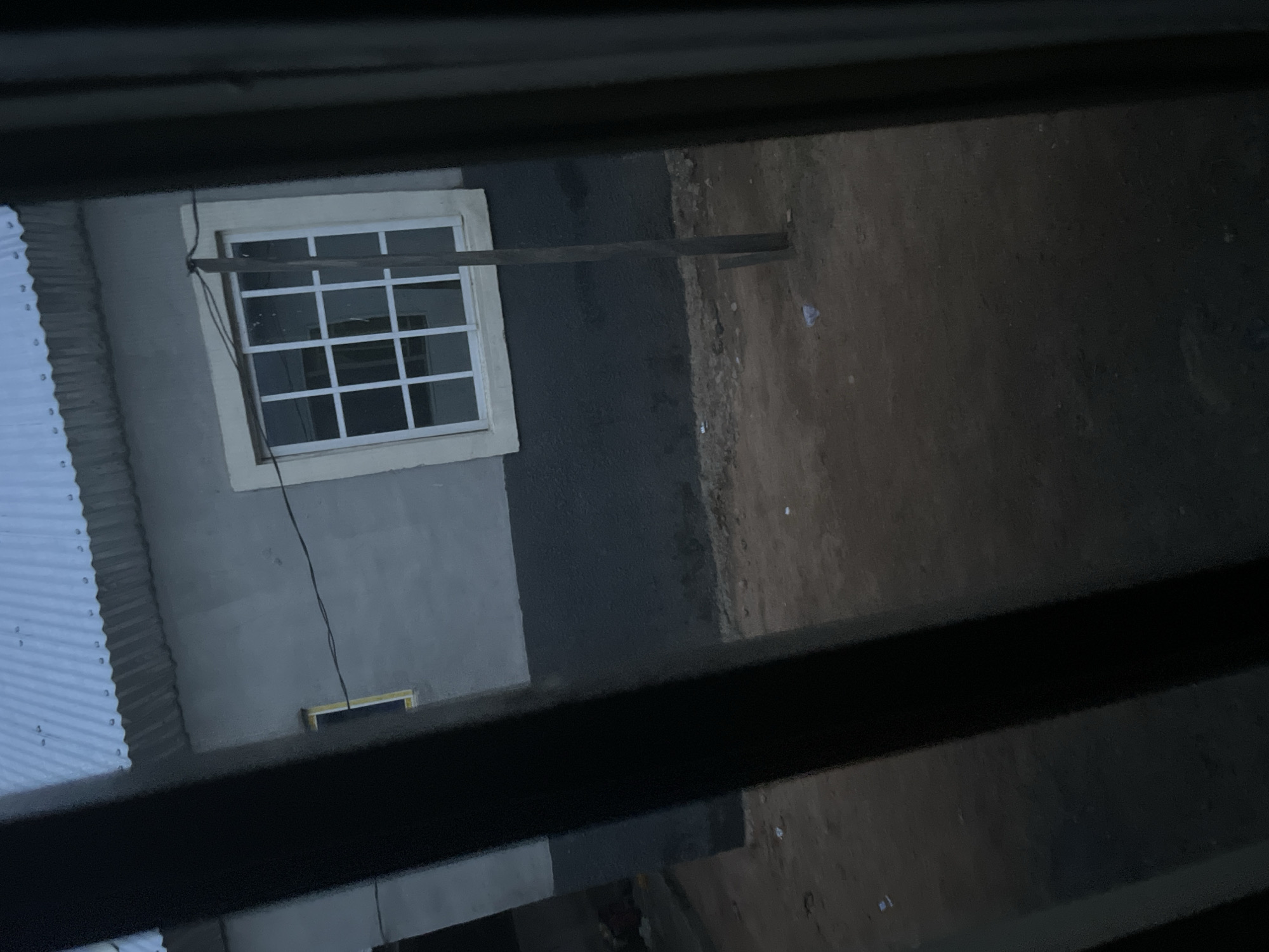 The image shows a building with a corrugated metal roof and a gray stucco exterior. There is a large window with a white grid pattern. A dark, rough surface runs along the base of the building. The ground in front of the building is composed of dirt and small pebbles. There are some pieces of litter scattered on the ground. A dark, vertical pole is positioned in front of the window. The scene appears to be outdoors in a relatively undeveloped area, possibly on the outskirts of Jalingo, Nigeria, given the context. The lighting suggests it is either early morning or late evening.