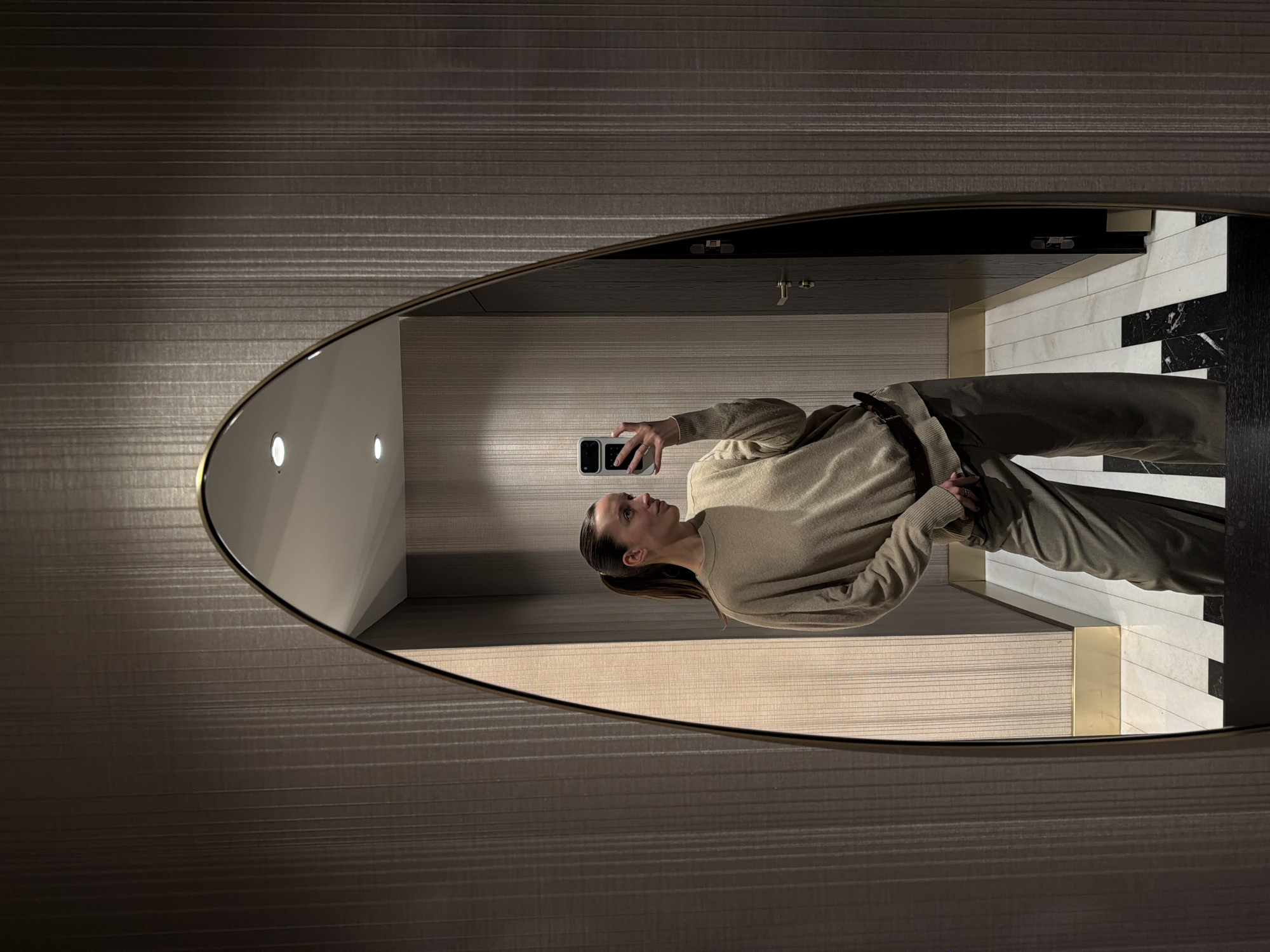 A woman is reflected in an oval mirror, holding a smartphone, in a bathroom with horizontal-striped walls. The mirror has a gold frame. The bathroom has black and white tile accents. The location is likely a hotel or apartment bathroom in Budva, Montenegro.