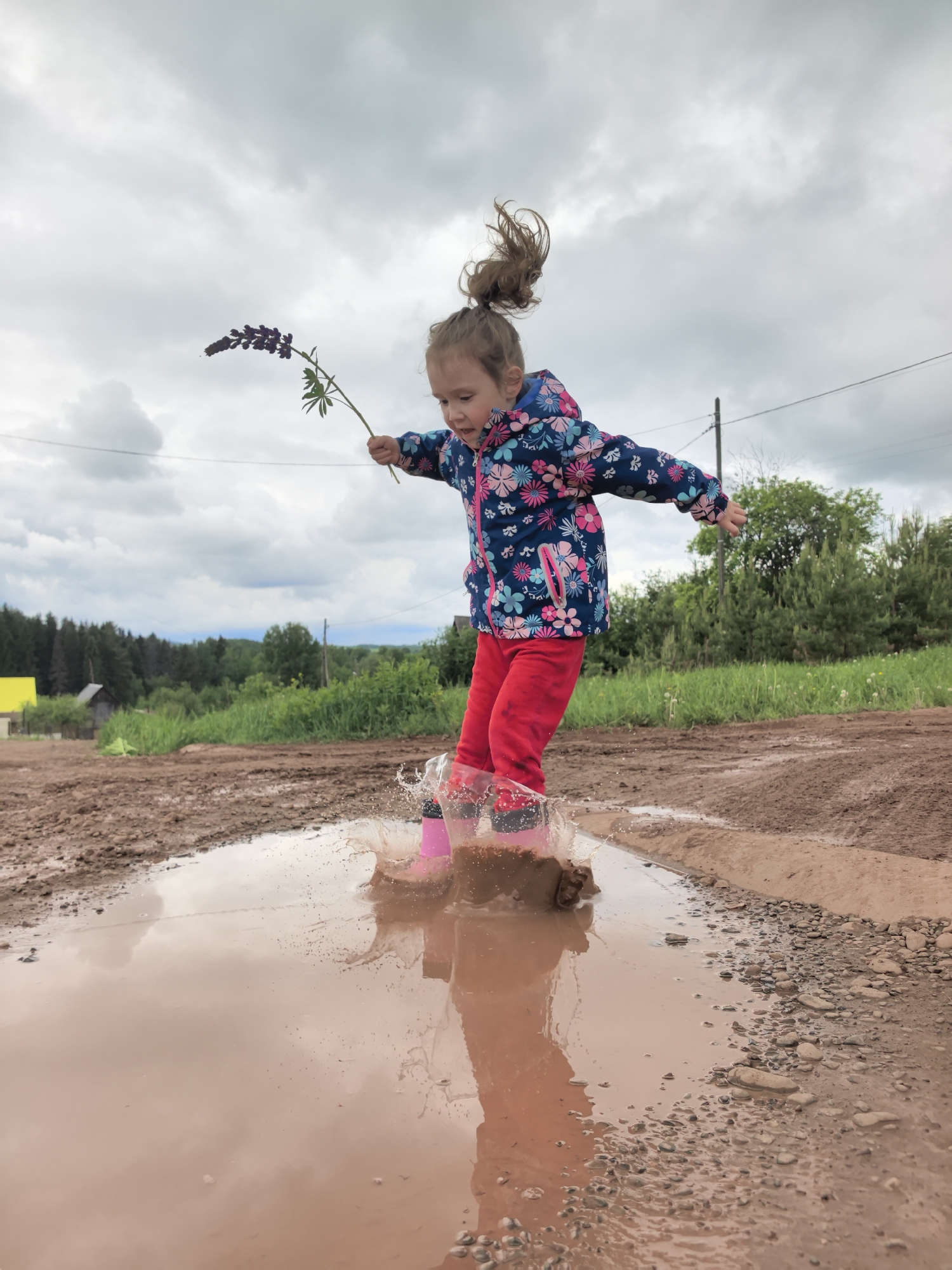 A young girl is joyfully jumping in a large puddle. She is wearing a blue floral jacket, red pants, and pink rubber boots. She is holding a purple flower in her right hand, and her arms are outstretched for balance. Her hair is pulled up in a high ponytail. The puddle is muddy brown, and the impact of her boots has created significant splashing. The surrounding ground is a muddy dirt road. The background features green trees, a small yellow building, and a cloudy sky. The location appears to be a rural or semi-rural area.