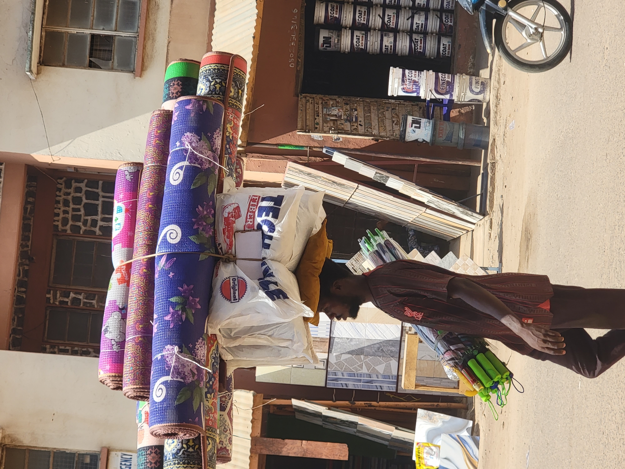 A man is seen on a street near Maiburiji in Nigeria. He is walking in the middle of the day and carrying an array of items on his head including rolled up mats in various designs, several white bags, a yellow pillow, and packages of green and blue handled items. He is wearing a maroon-colored top and brown trousers. The background includes shop exteriors with stacked buckets of paint, a motorcycle, and building facades. A sign on a bucket of paint says "FINECOAT", and another white bag says "original FIBER". A phone number "0802553166" is visible above one of the shops. Tiles and construction materials appear in front of the shops.