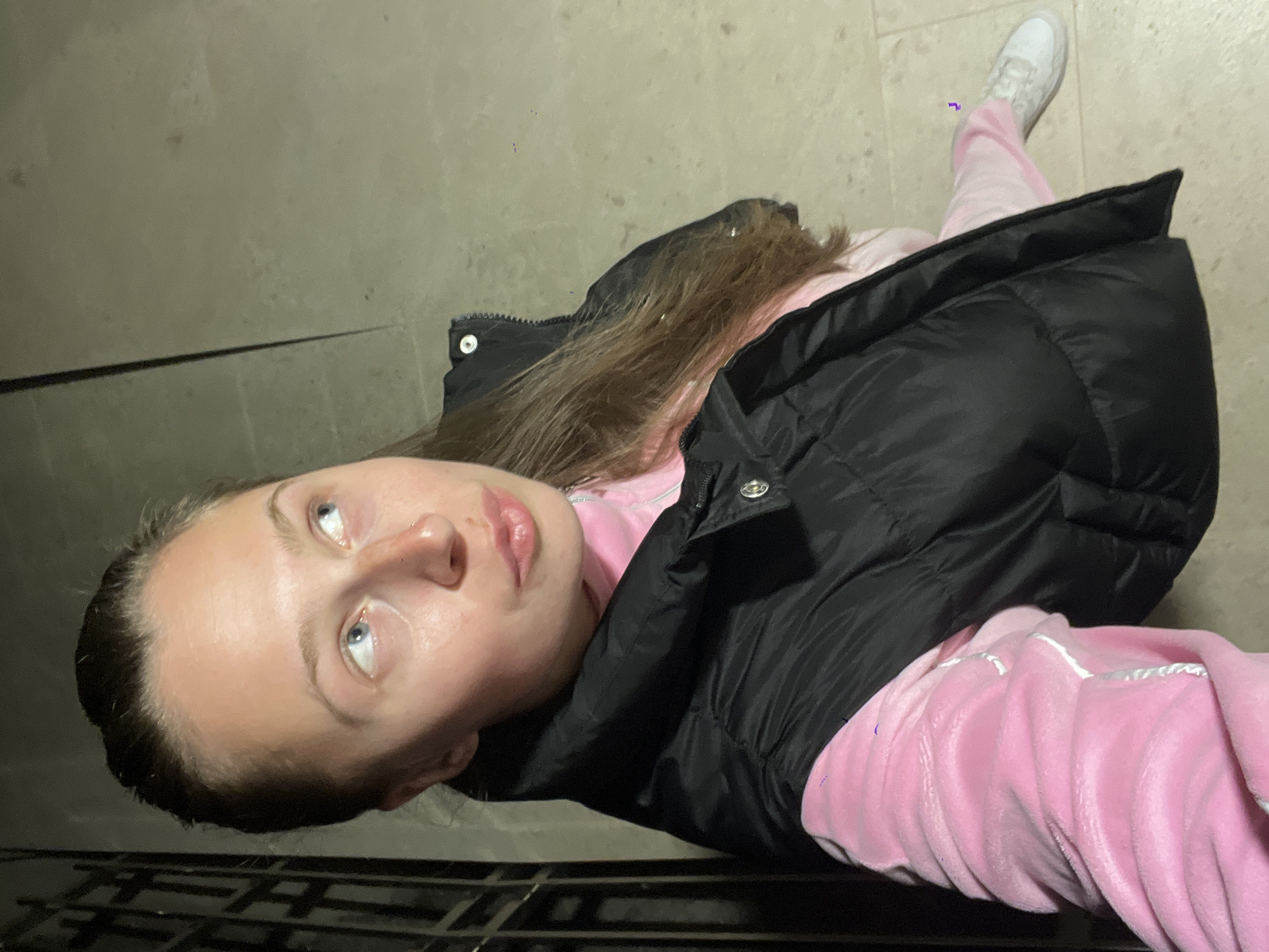 A young woman is standing outdoors on a stone surface. She is wearing a pink tracksuit and a black vest. The woman has blonde hair and blue eyes. Her feet are visible and she is wearing white sneakers. The location appears to be an outdoor, paved area with a metal grate visible at the bottom of the frame. Bečići, Montenegro is not discernable from the photograph.