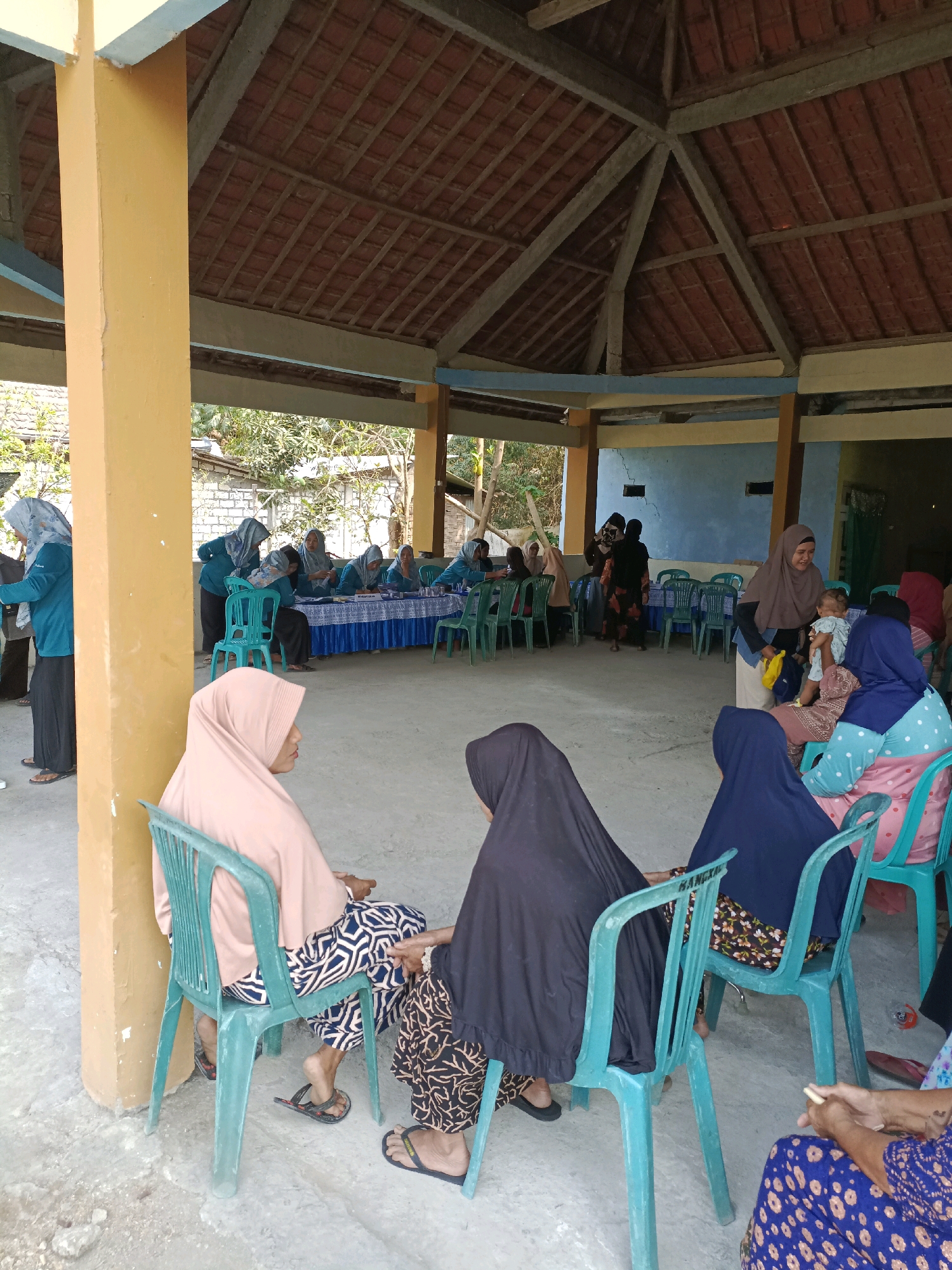 In Rancang Kencono, Indonesia, a group of women, most wearing hijabs, are gathered under an open-sided structure with a tiled roof and a supporting yellow pillar. Some women are seated on teal plastic chairs in the foreground, facing towards a table area in the background. Others are standing or seated around the table, which is covered with a blue and white patterned cloth. Several women appear to be engaged in a meeting or discussion, with some looking at documents. One woman in the background is holding a baby. The environment is outdoors with visible trees and buildings in the distance. The ground is concrete.