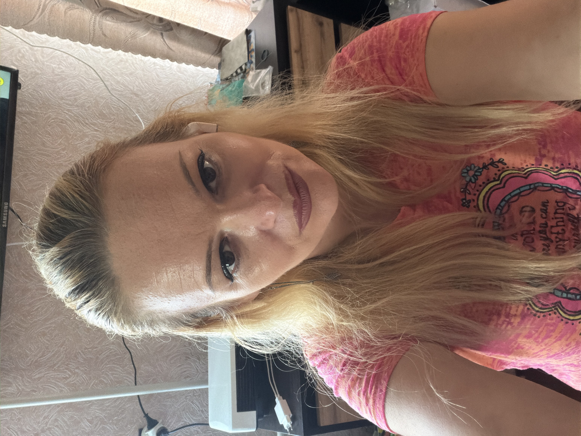 A medium shot selfie shows a Caucasian woman with blonde hair and fair skin, wearing a pink t-shirt. She is indoors, with a blurred background that includes a white electronic device, a dark desk with various objects, and textured wallpaper. Her hair is pulled back, and she is wearing an earbud in her right ear. The lighting suggests an indoor environment, possibly a home office or study. No specific details indicate the city of Chernihiv, Ukraine.