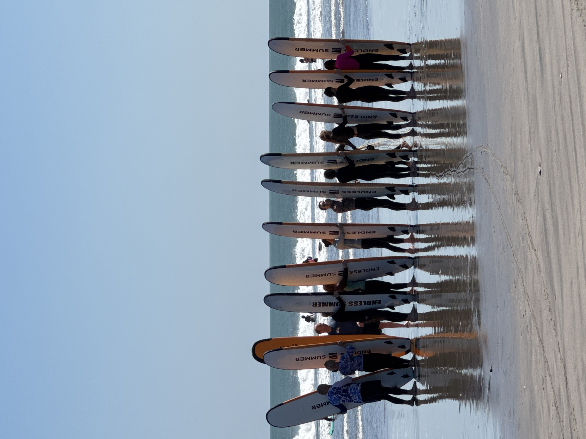 A group of approximately 15 individuals, some in wetsuits and others in casual attire, stand on a wet sandy beach, holding upright surfboards. The surfboards, some with "ENDLESS SUMMER" branding, are arranged in a line. The ocean, with gentle waves breaking, is visible behind them, and the sky is clear and blue. The scene suggests a surfing lesson or group activity. The location is Kuta, Indonesia.